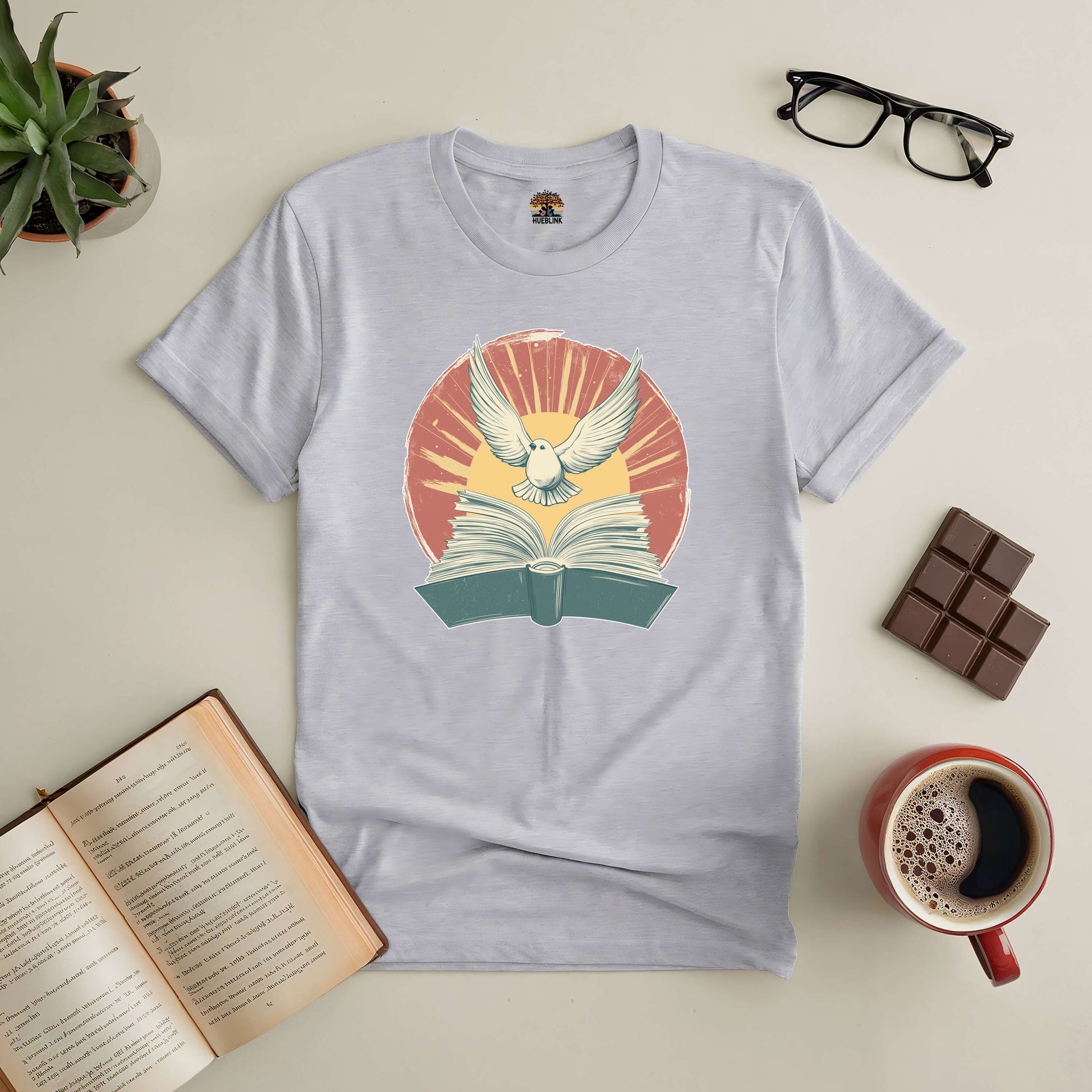 Boundless Horizons Tee featuring open book and bird design, surrounded by coffee, book, glasses, chocolate, and plant on a table.