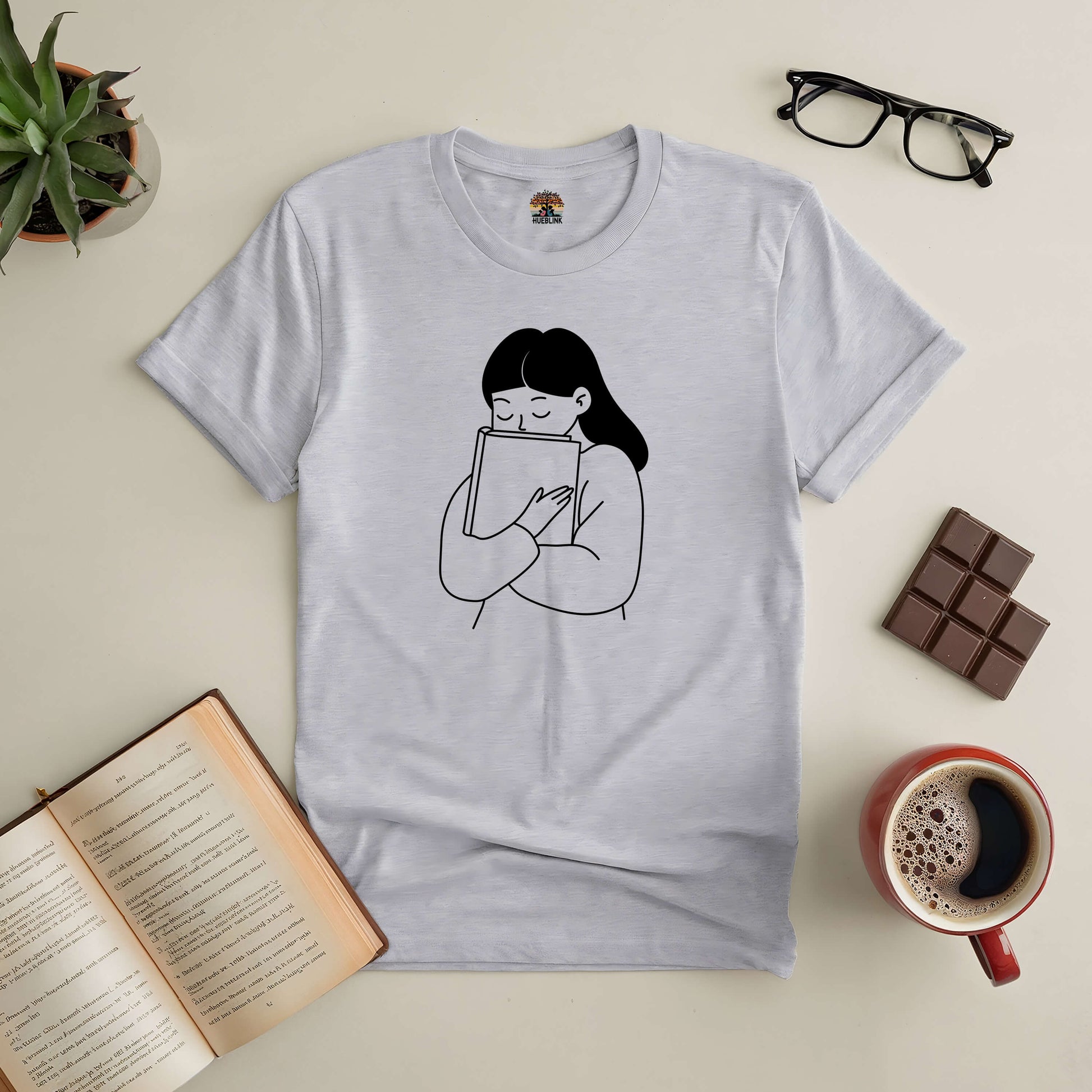 Minimalistic tee for book lovers featuring a line drawing of a woman hugging a book, surrounded by coffee, chocolate, and reading glasses.