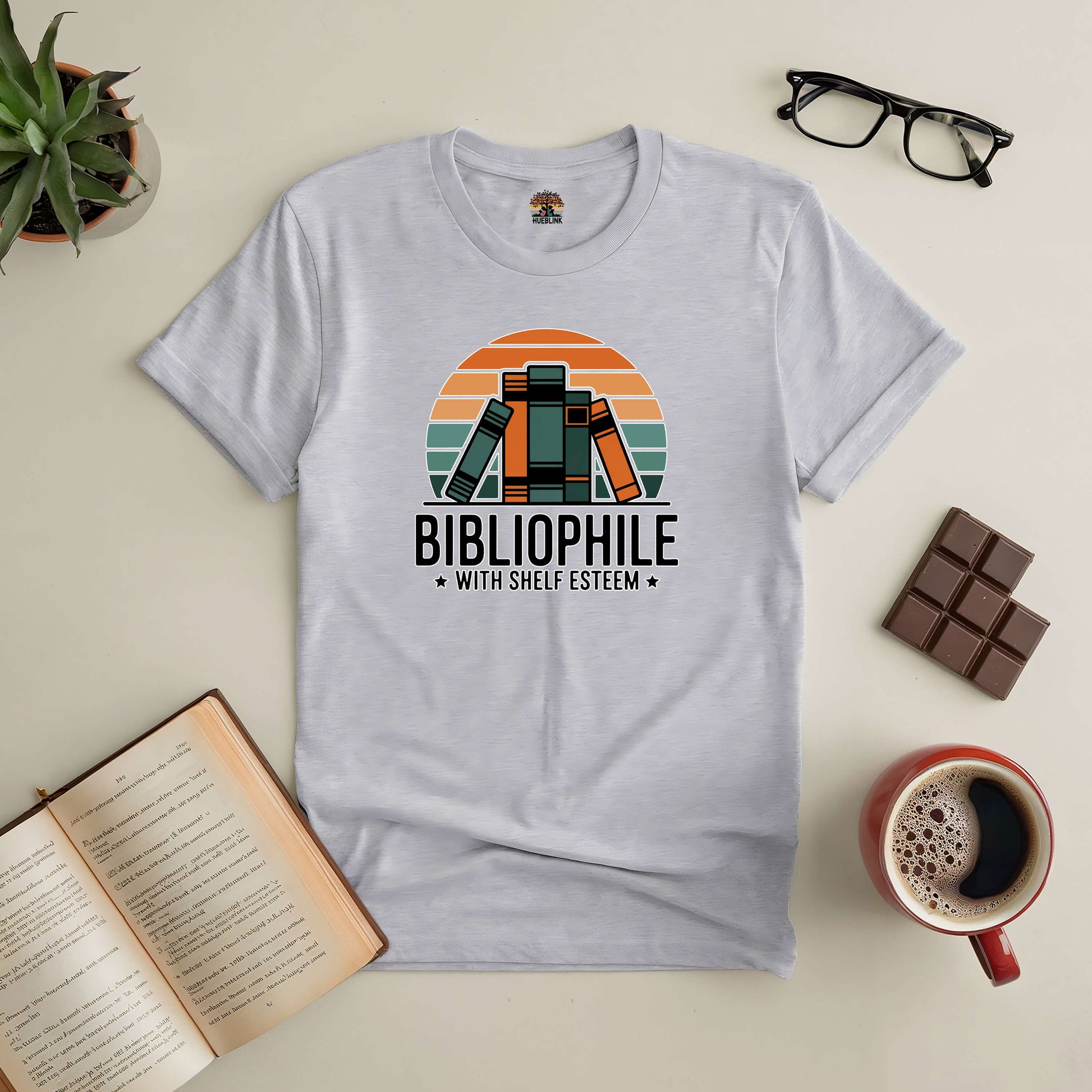 Retro-style "Bibliophile with Shelf Esteem" tee, perfect for book lovers, displayed with coffee, glasses, and an open book.