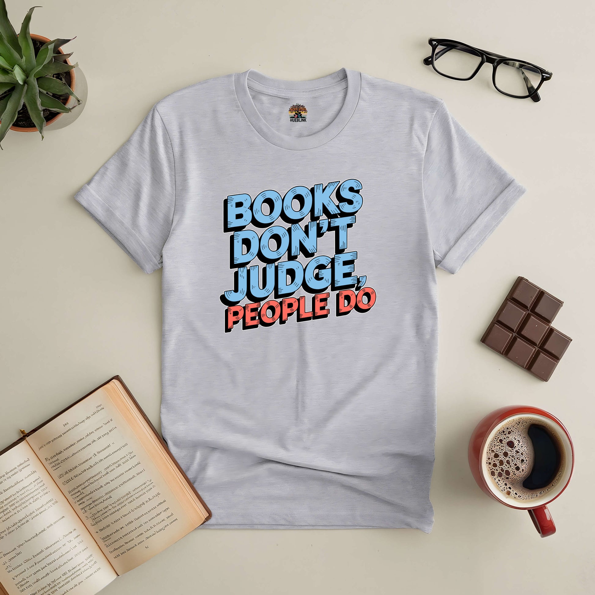 Grey tee with "Books Don't Judge, People Do" text, surrounded by glasses, chocolate, coffee, and an open book on a table.