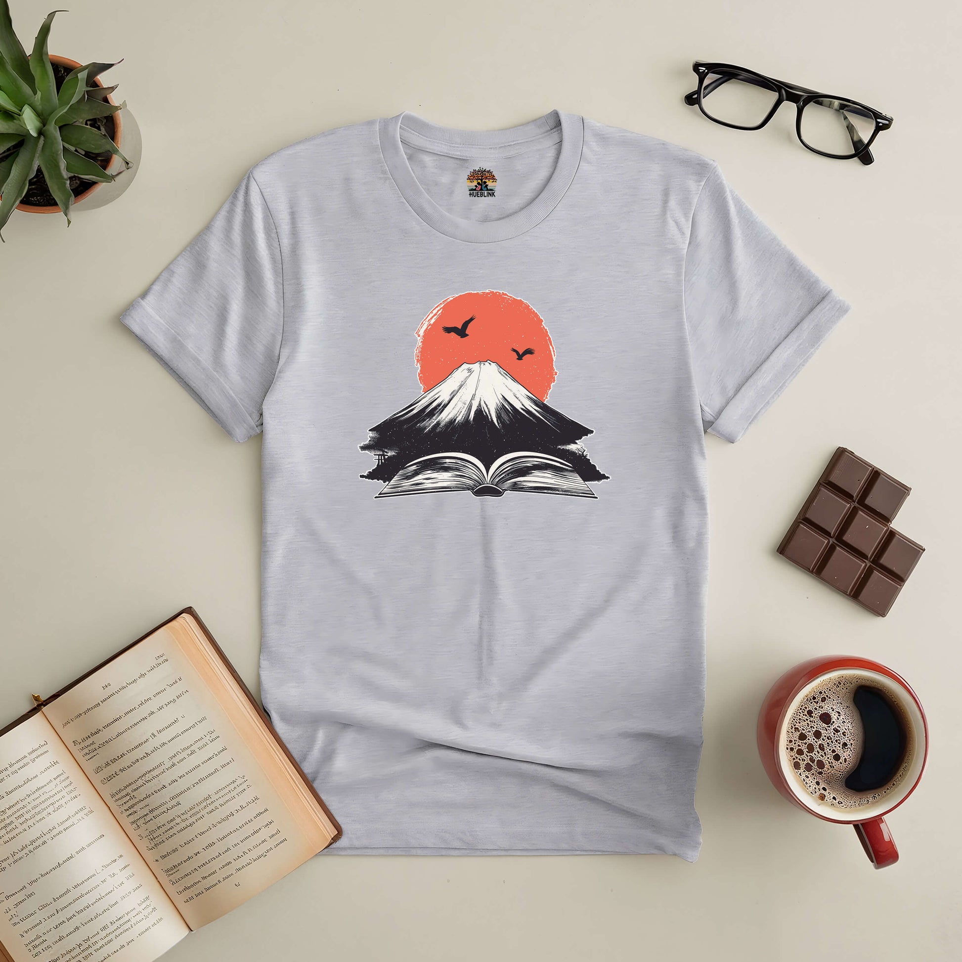 Eruption of Imagination Tee with open book design, symbolizing creative adventure, surrounded by coffee, glasses, and chocolate.