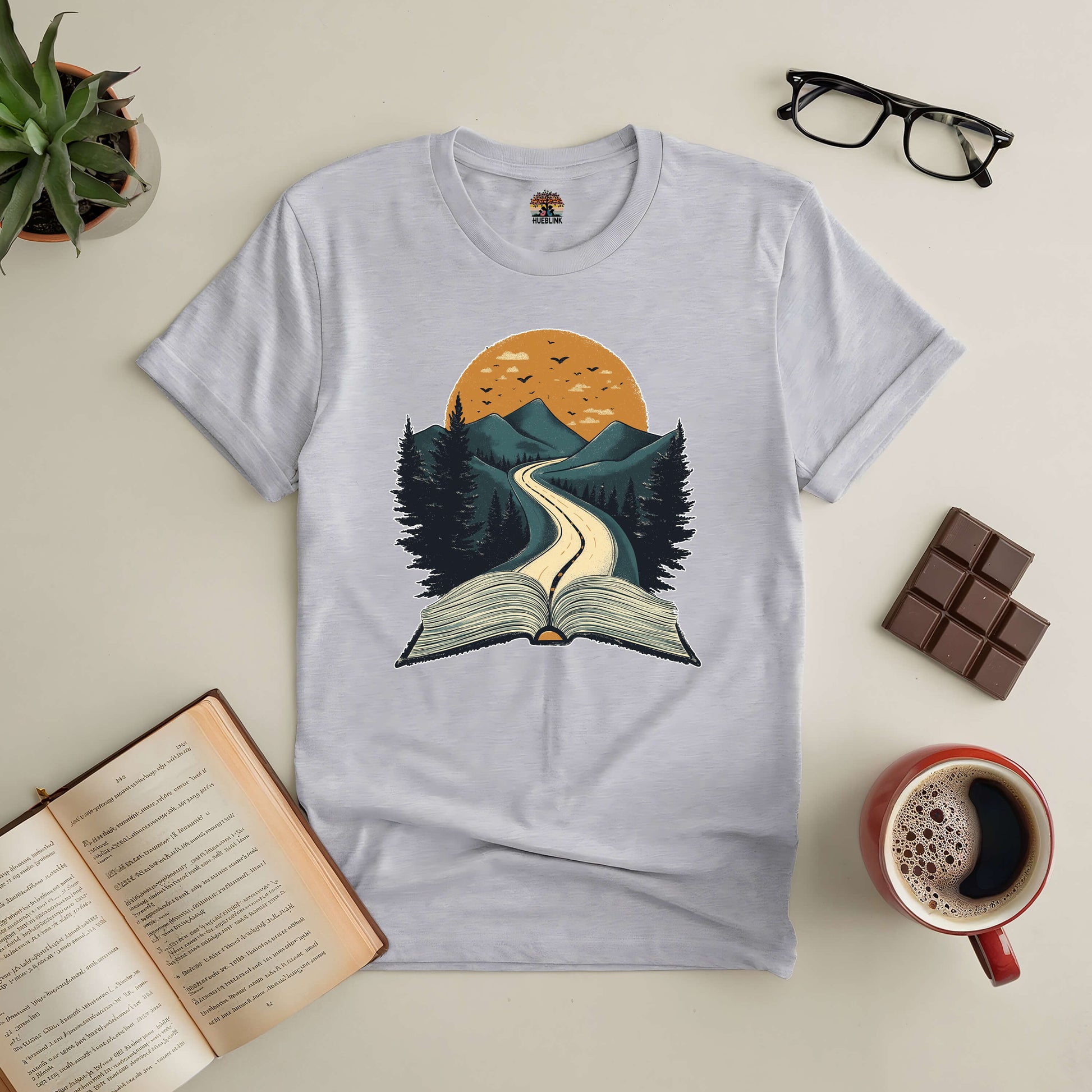 Starting Point Tee with book and road design, surrounded by coffee, glasses, open book, and chocolate on a table. Perfect for readers.