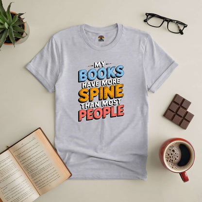 White tee with "My Books Have More Spine Than Most People" design, surrounded by open book, chocolate, glasses, and coffee.