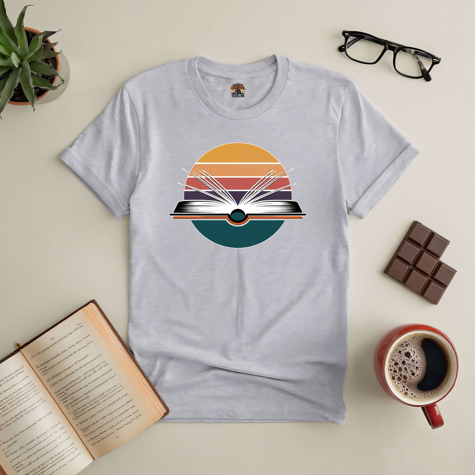 Gateway to New Horizons Tee with book graphic, surrounded by coffee, chocolate, glasses, and an open book.