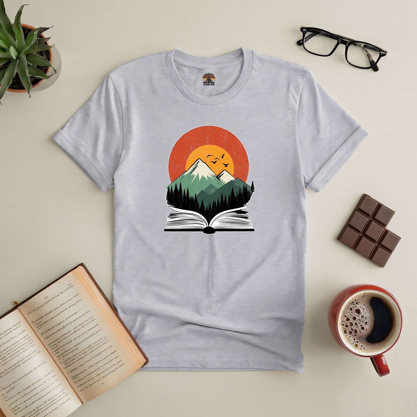 "Mountains of Imagination Tee depicting a mountain scene emerging from an open book, surrounded by coffee, glasses, and chocolate"