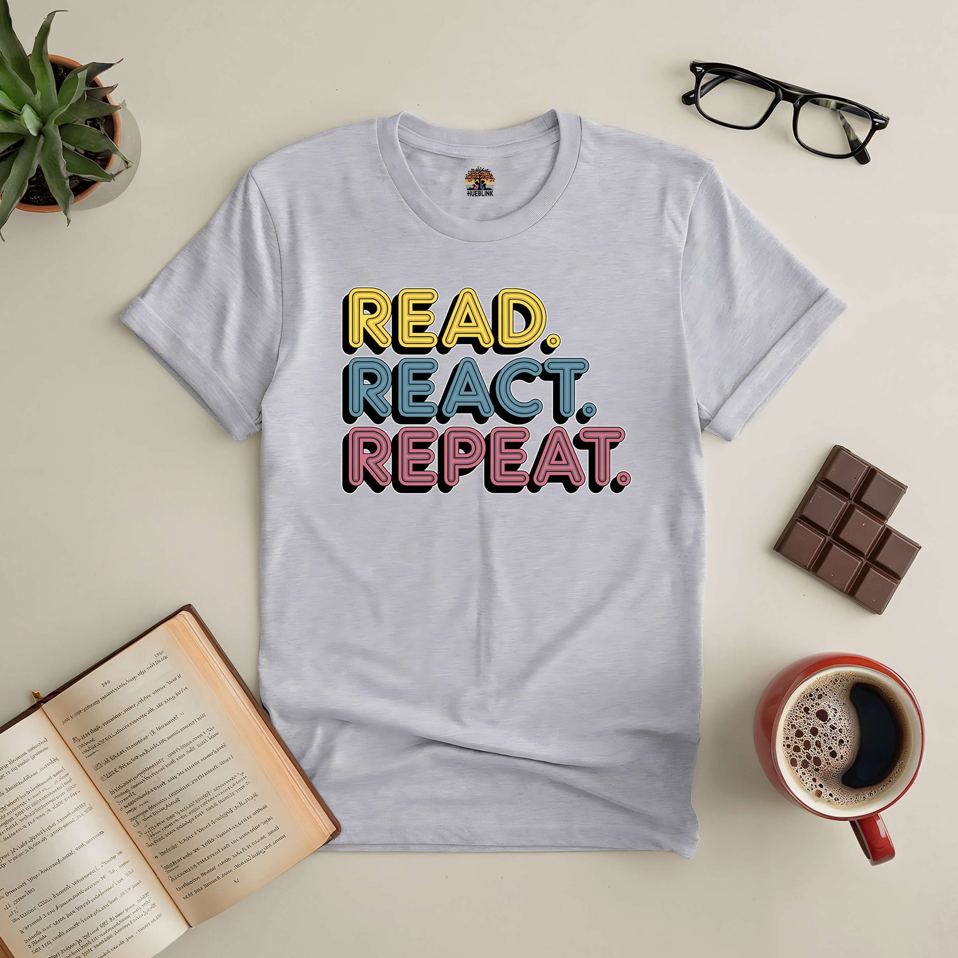 "Read. React. Repeat. Tee with colorful text surrounded by book, coffee, glasses, and chocolate on a cozy table setup."