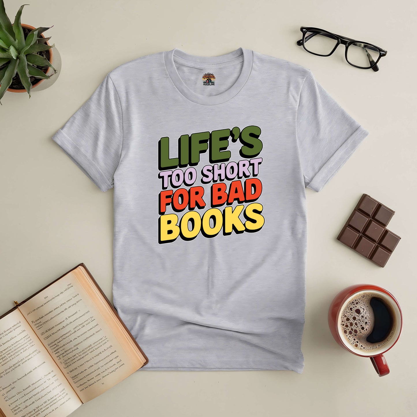 Life's Too Short for Bad Books Tee surrounded by open book, coffee, chocolate, and glasses on a table. Perfect for book lovers.