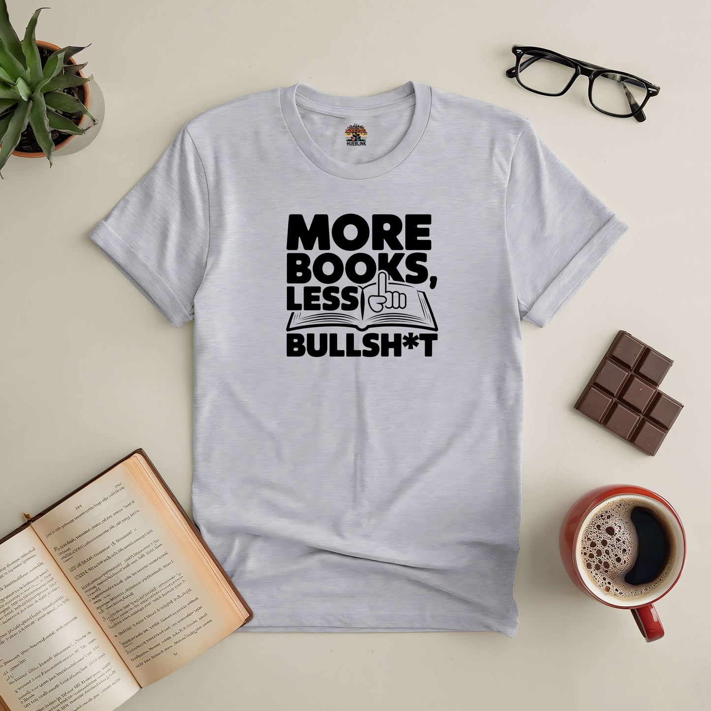 Gray More Books, Less Bullsh*t Tee surrounded by an open book, glasses, chocolate, coffee, and a potted plant.