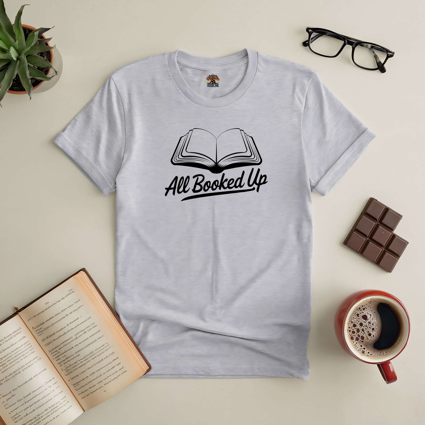 "All Booked Up Tee featuring an open book design, surrounded by coffee, an open book, chocolate, glasses, and a plant."