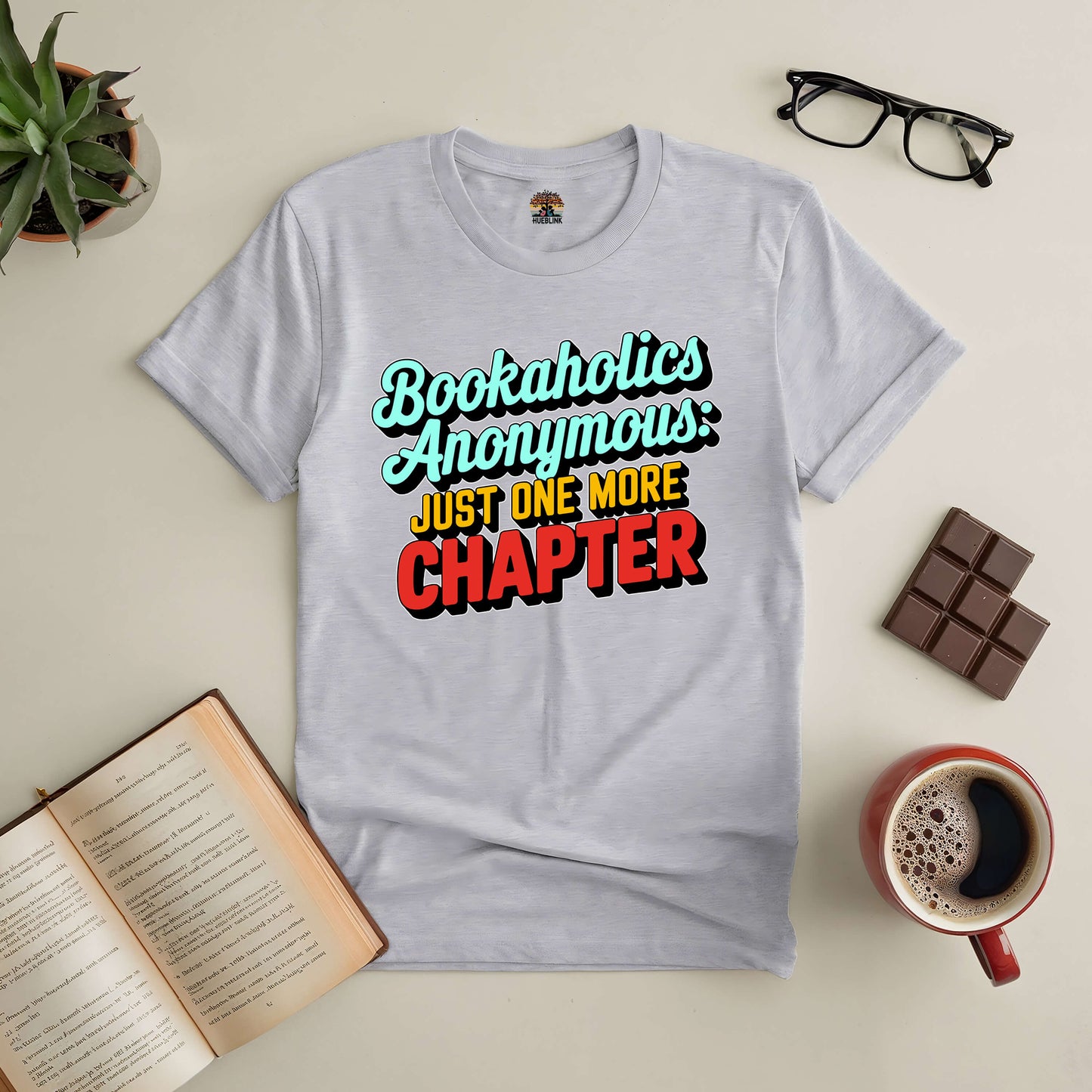 Gray "Bookaholics Anonymous: Just One More Chapter" tee surrounded by glasses, book, chocolate, plant, and coffee.