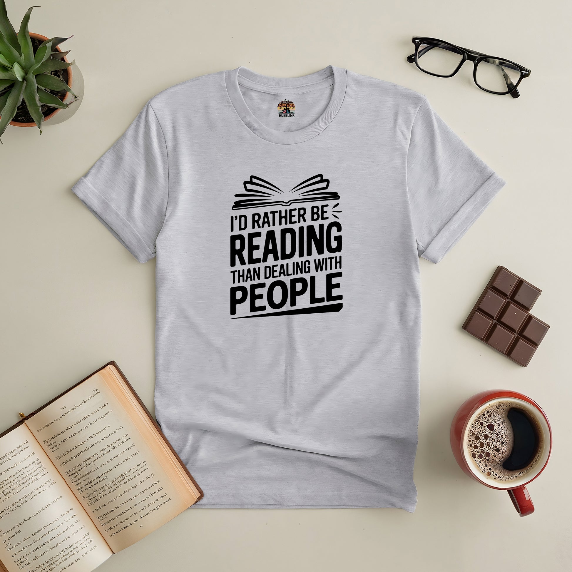 Gray tee with "I'd Rather Be Reading Than Dealing with People" text, surrounded by glasses, open book, chocolate, coffee, and plant.