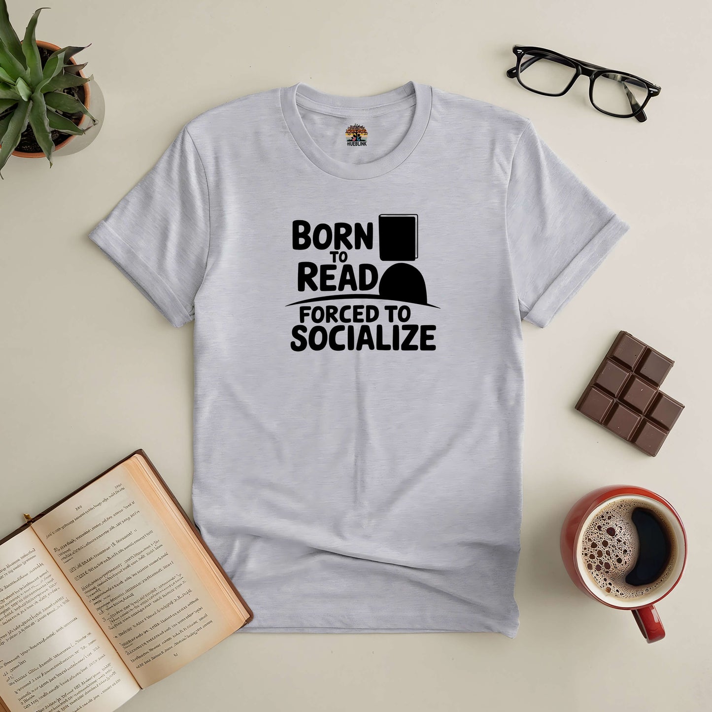 "Born to Read, Forced to Socialize Tee for introverts with book, coffee, chocolate, glasses, and plant on a table"