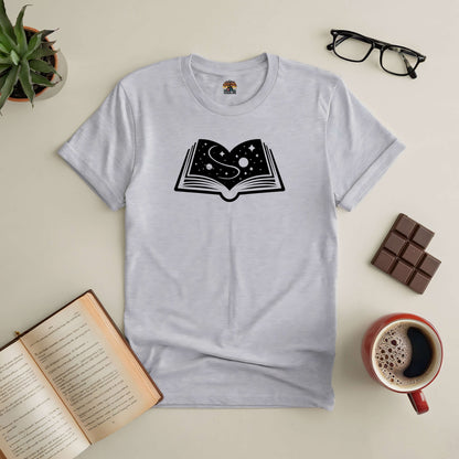 Gray t-shirt with illustrated open book and galaxy design, surrounded by glasses, chocolate, coffee, and open book on table.