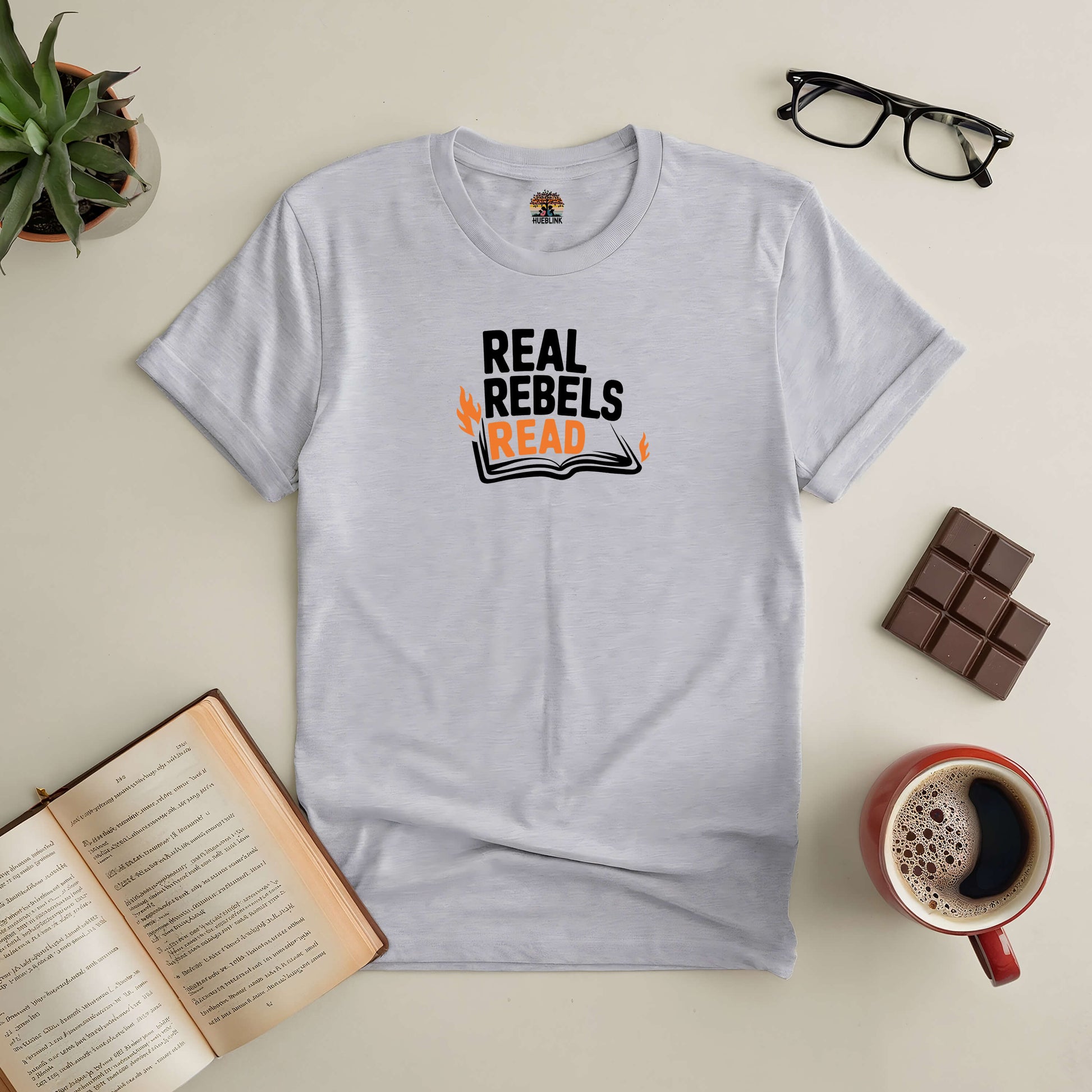 White "Real Rebels Read" t-shirt displayed with open book, glasses, chocolate, and coffee on a table.