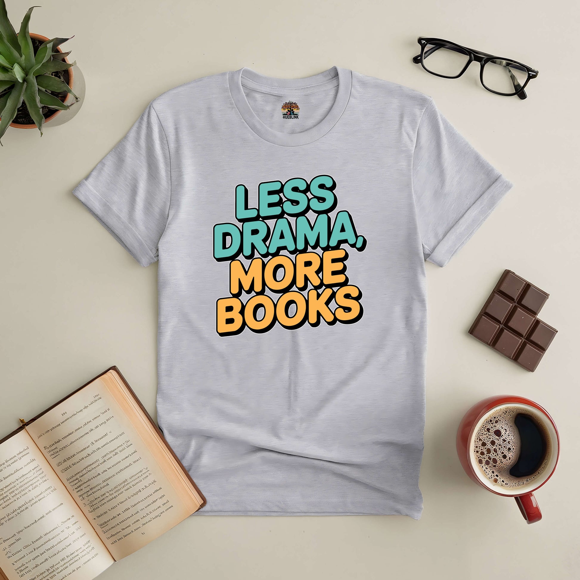 Grey tee with "Less Drama, More Books" text, surrounded by an open book, glasses, chocolate, and coffee on a table.