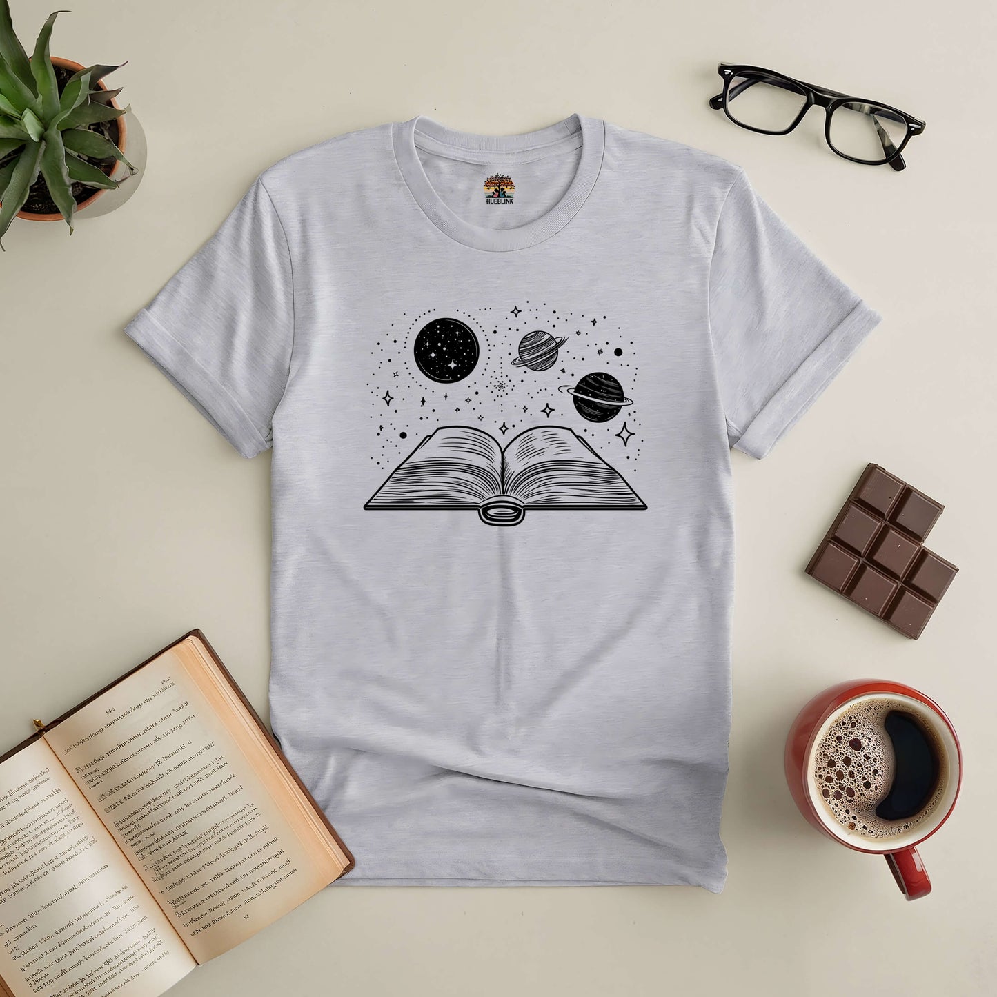 Space-themed t-shirt with an open book, planets, and stars, surrounded by a coffee cup, chocolate, and reading glasses.