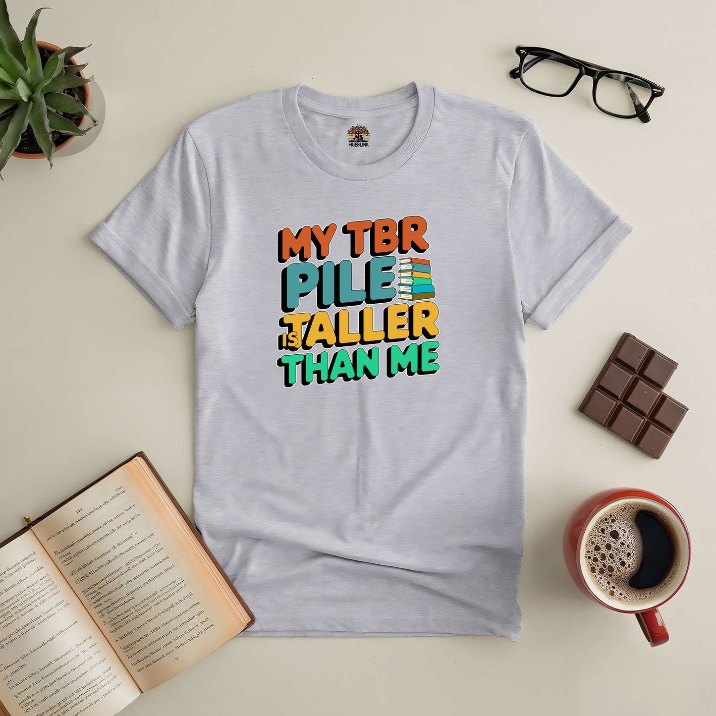 Grey tee with "My TBR Pile is Taller Than Me" text, surrounded by glasses, coffee, chocolate, and an open book. Perfect for book lovers.