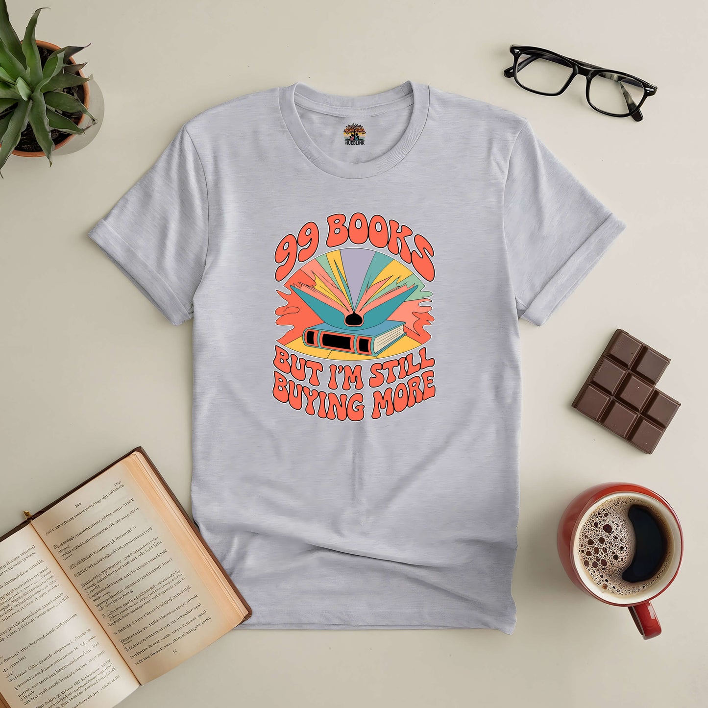 Gray "99 Books and Counting!" tee showcasing a colorful book design, surrounded by glasses, chocolate, coffee, and an open book.