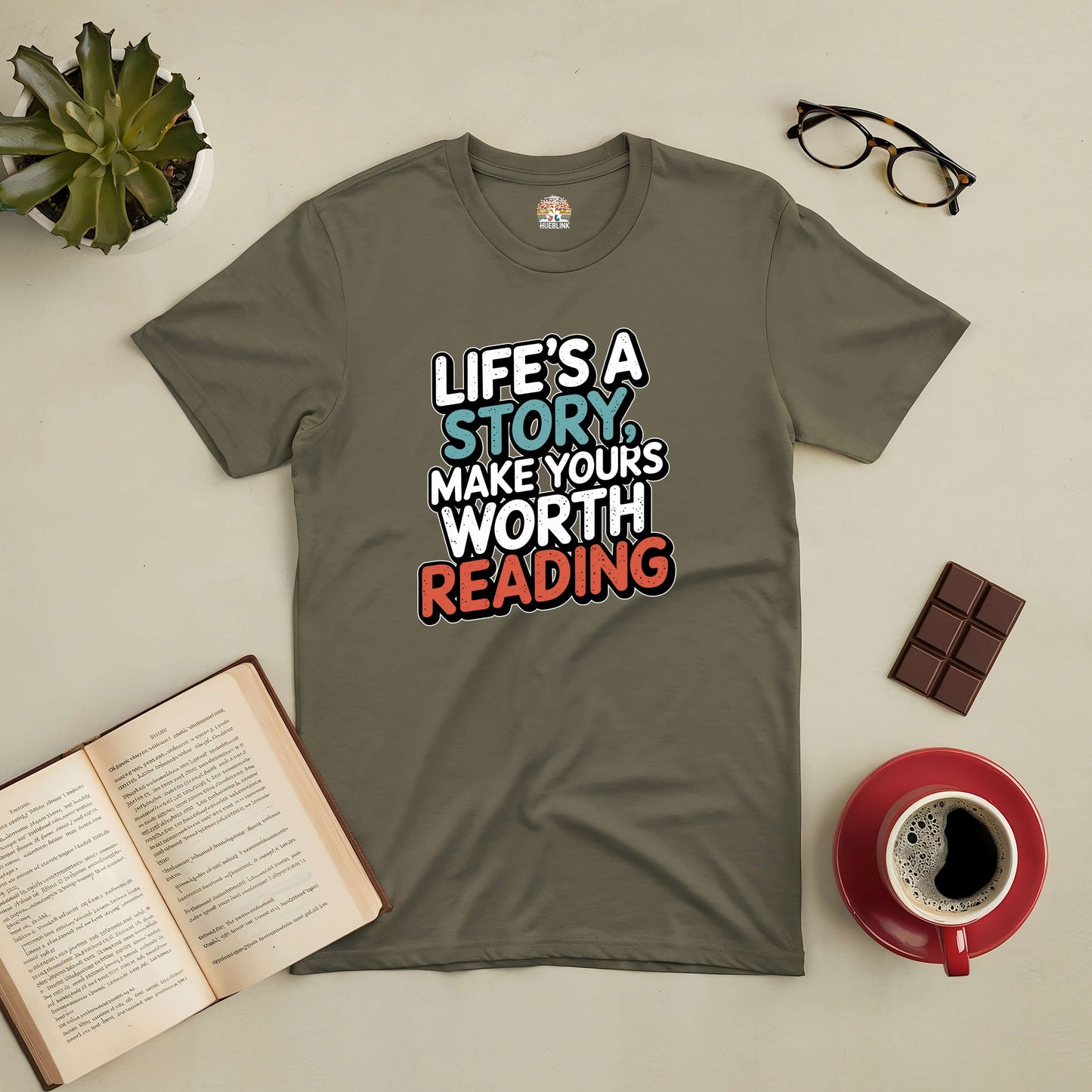 Motivational tee saying "Life's a Story, Make Yours Worth Reading," surrounded by coffee, book, glasses, and chocolate.