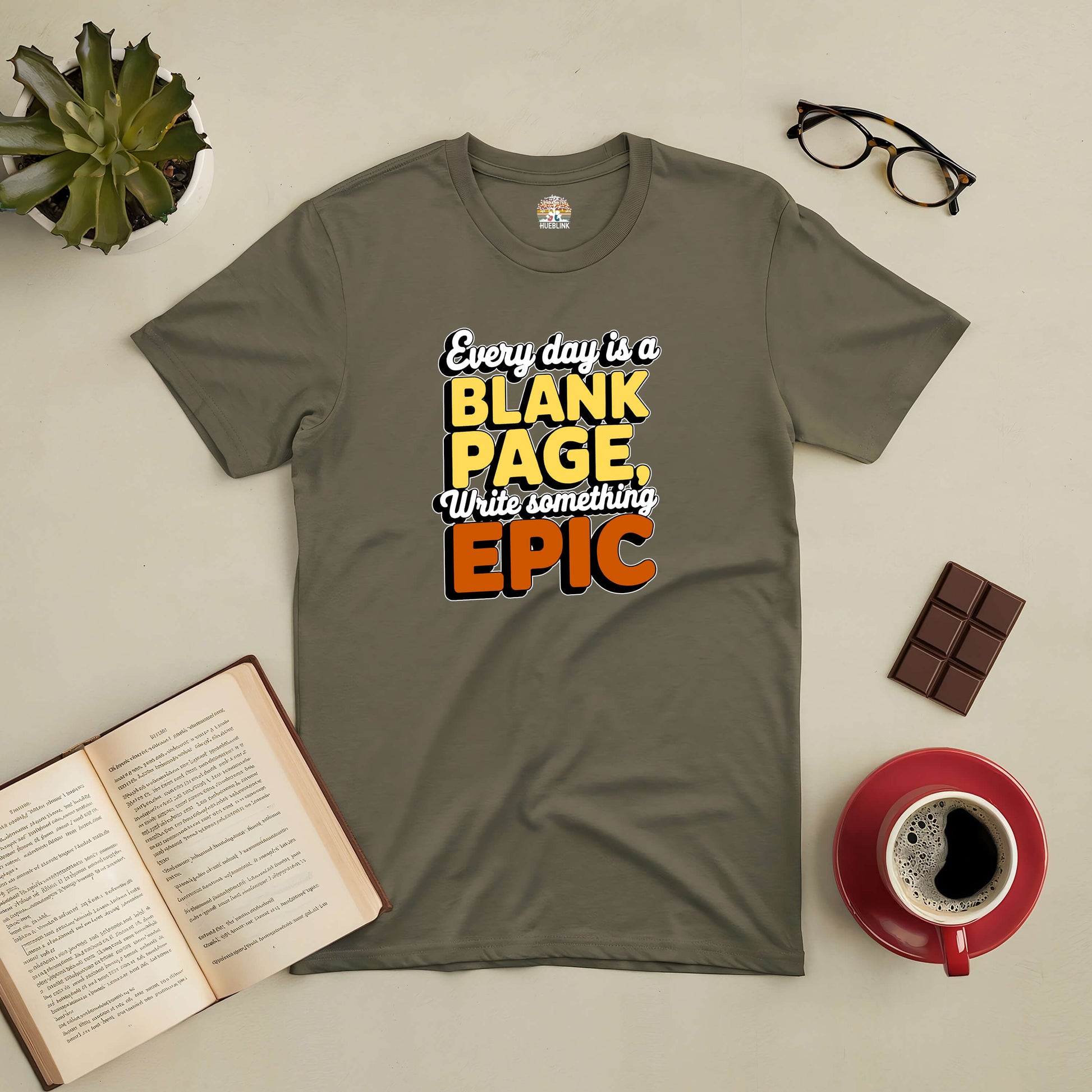 Inspirational tee with "Every Day is a Blank Page, Write Something Epic" design beside open book, coffee, and glasses.