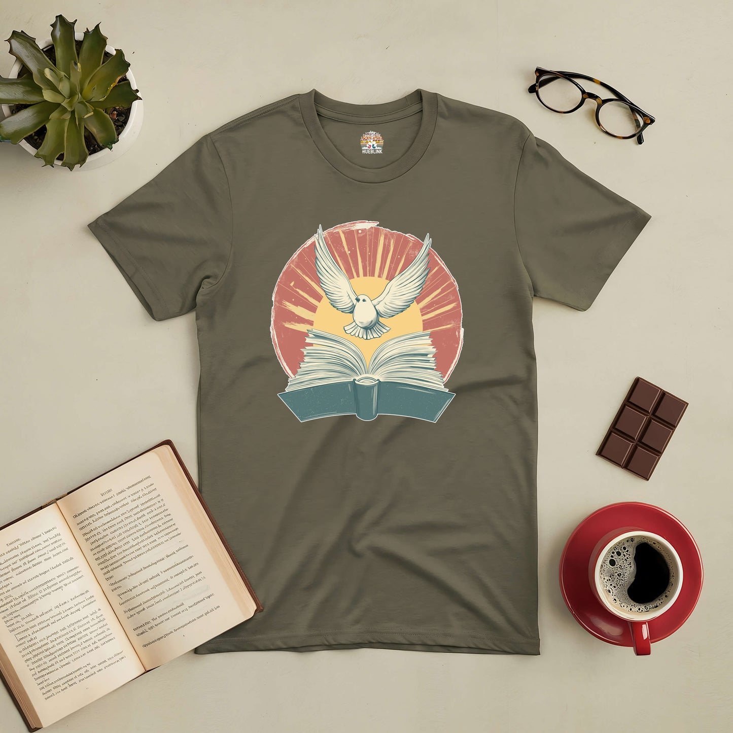 "Boundless Horizons Tee with an open book and flying bird design, surrounded by a coffee cup, glasses, and succulent"