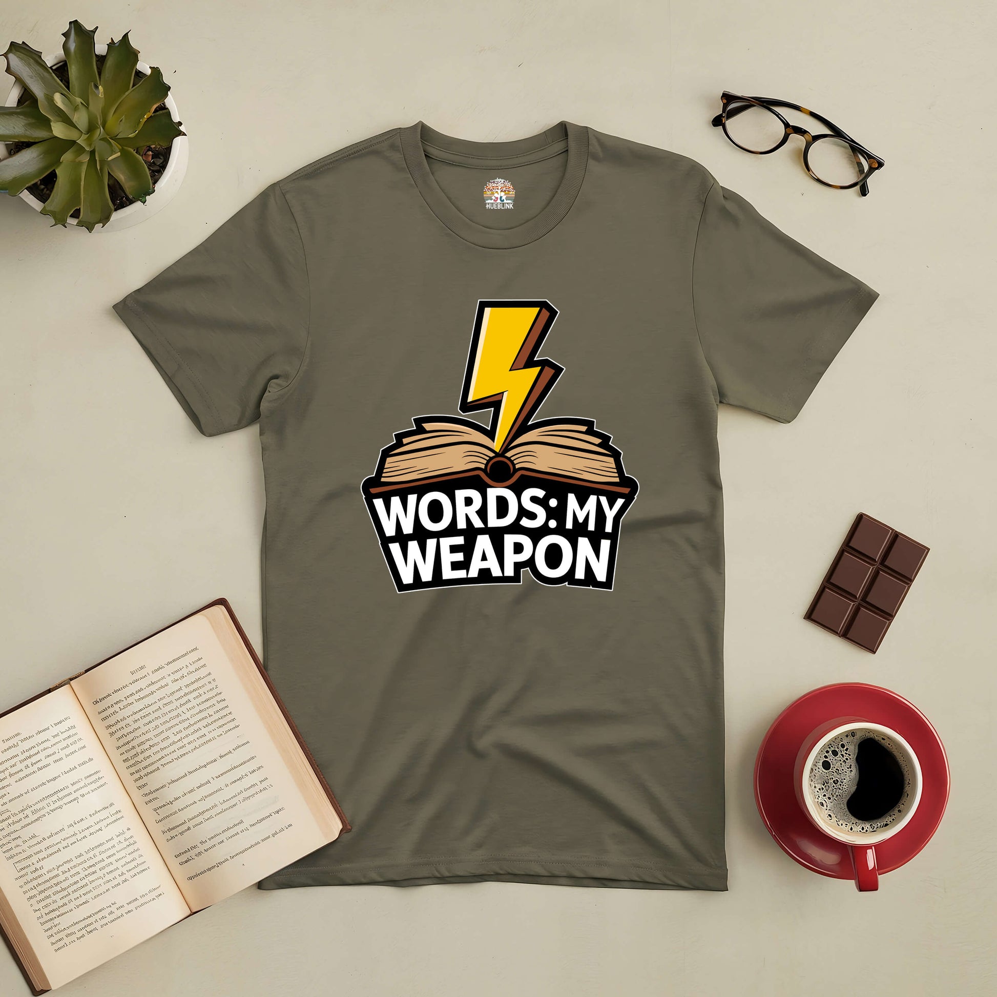 "Words: My Weapon Tee with book and lightning design for language enthusiasts on table with coffee, chocolate, and open book."