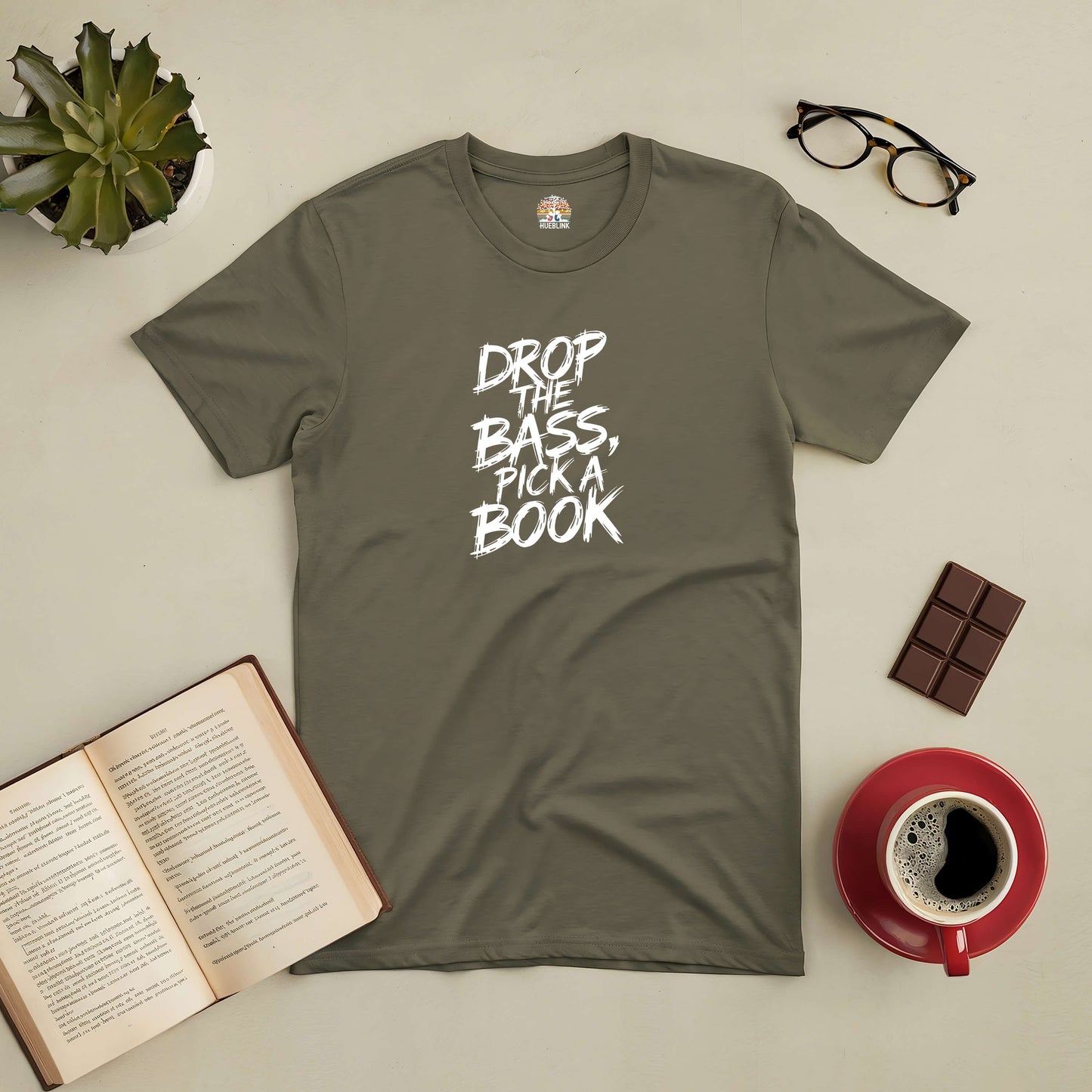 Olive green tee with "Drop The Bass, Pick a Book" print, surrounded by open book, coffee, glasses, and chocolate.