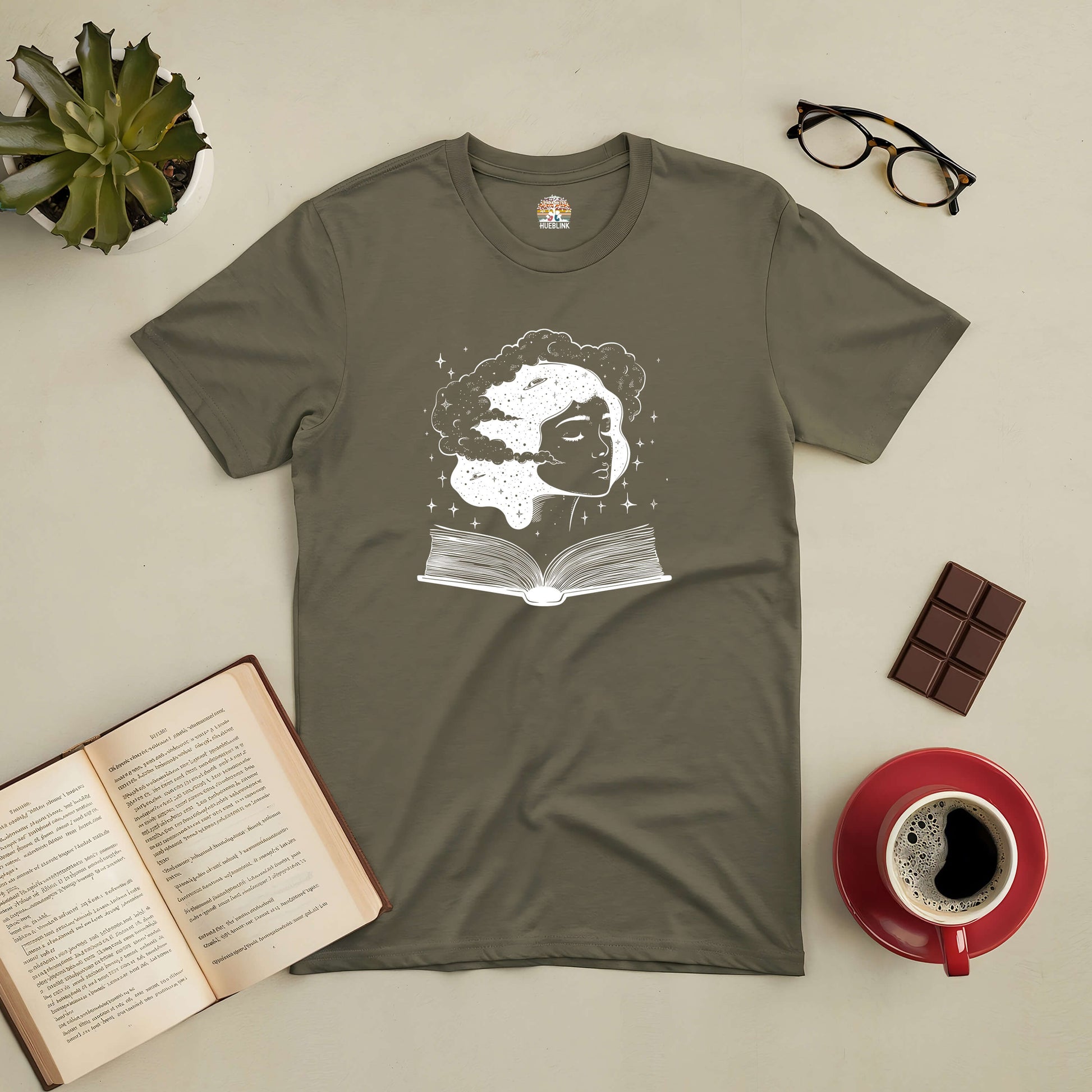 Universal Consciousness Tee with cosmic book design, surrounded by coffee, glasses, chocolate, and an open book.