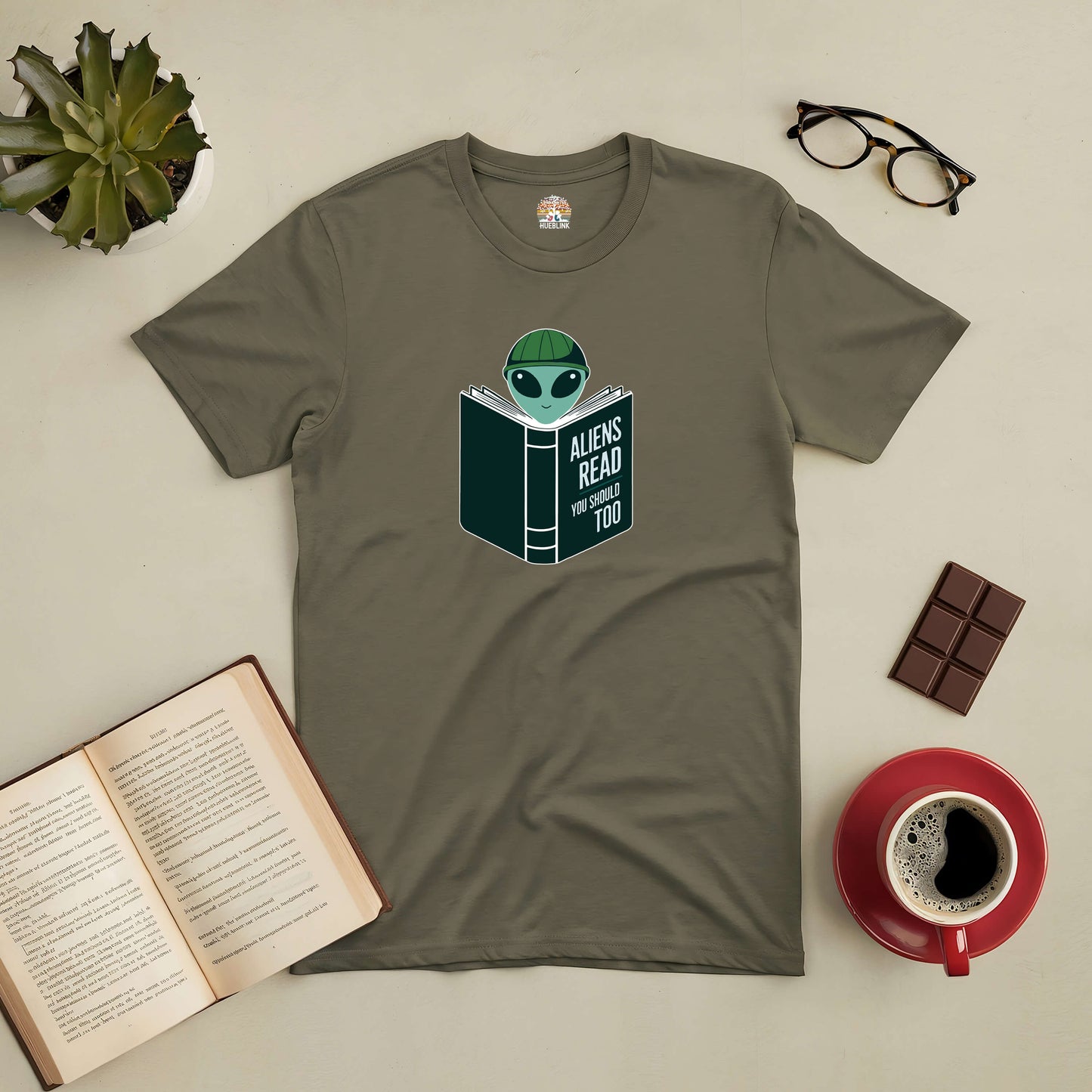 Intergalactic Habit Tee featuring an alien reading a book, ideal for book lovers and sci-fi fans, surrounded by coffee and books.