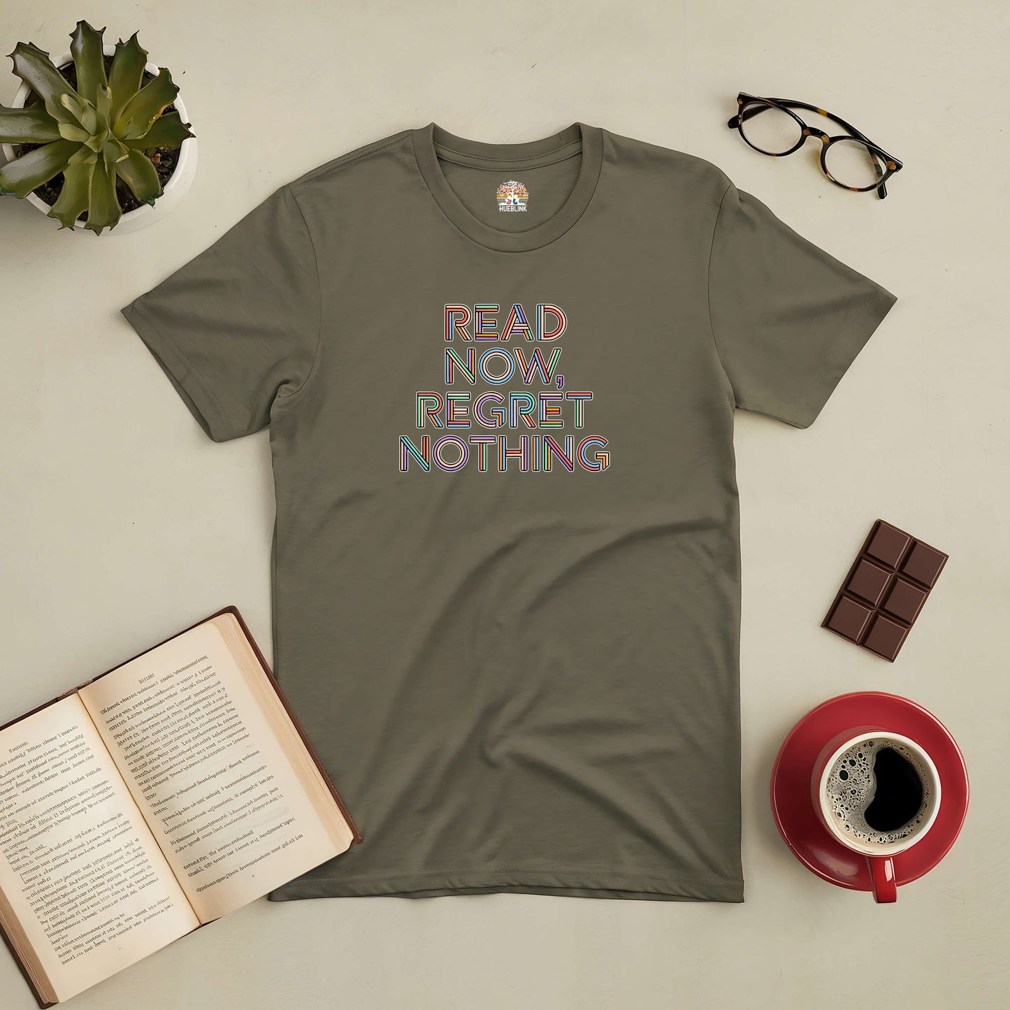 Olive green "Read Now, Regret Nothing" tee with a book, coffee, glasses, and chocolate on a desk, inspiring reading adventure.