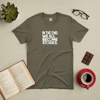 Olive green tee with "In The End, We All Become Stories" text, surrounded by a plant, glasses, chocolate, book, and coffee cup.