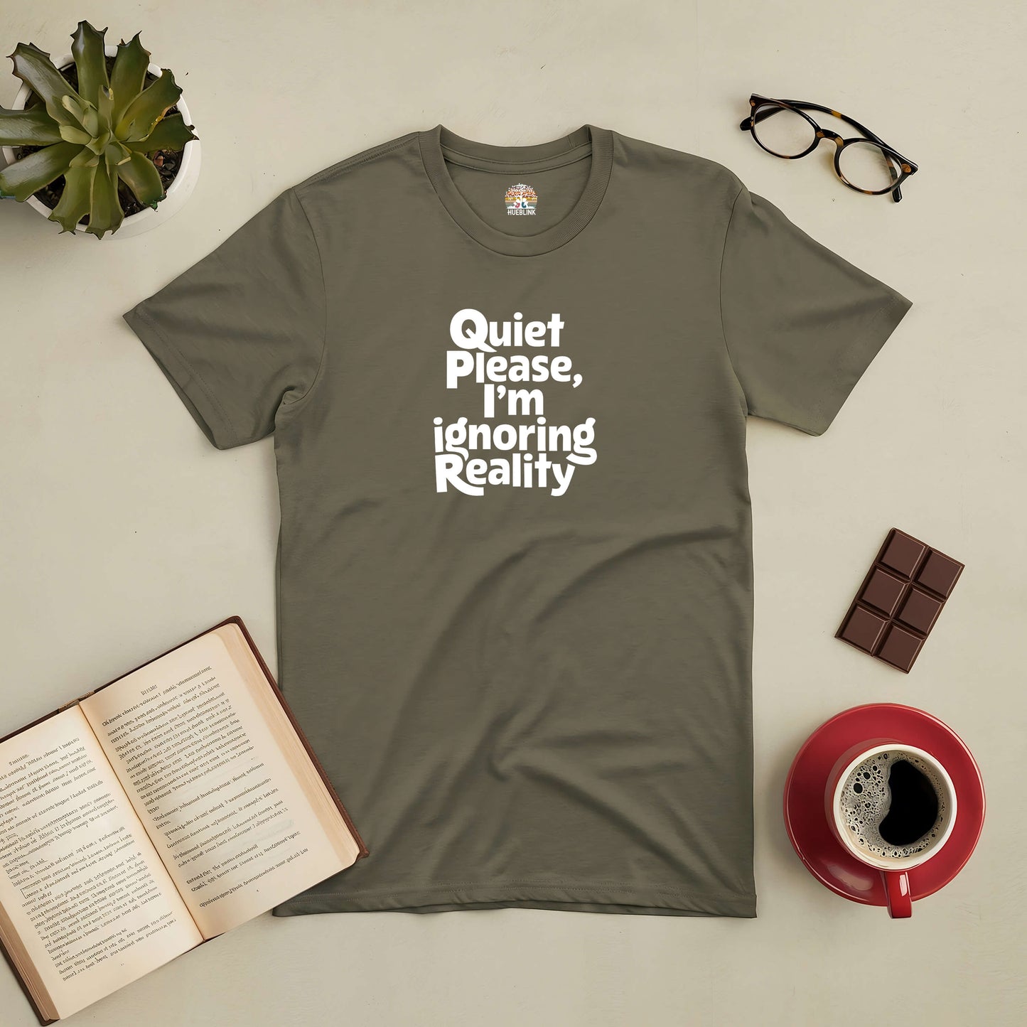 Green "Quiet Please, I'm Ignoring Reality" tee surrounded by a book, coffee, chocolate, glasses, and a succulent plant.