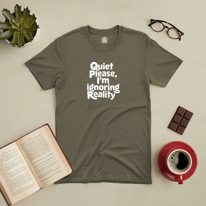 Green "Quiet Please, I'm Ignoring Reality" tee surrounded by a book, coffee, chocolate, glasses, and a succulent plant.