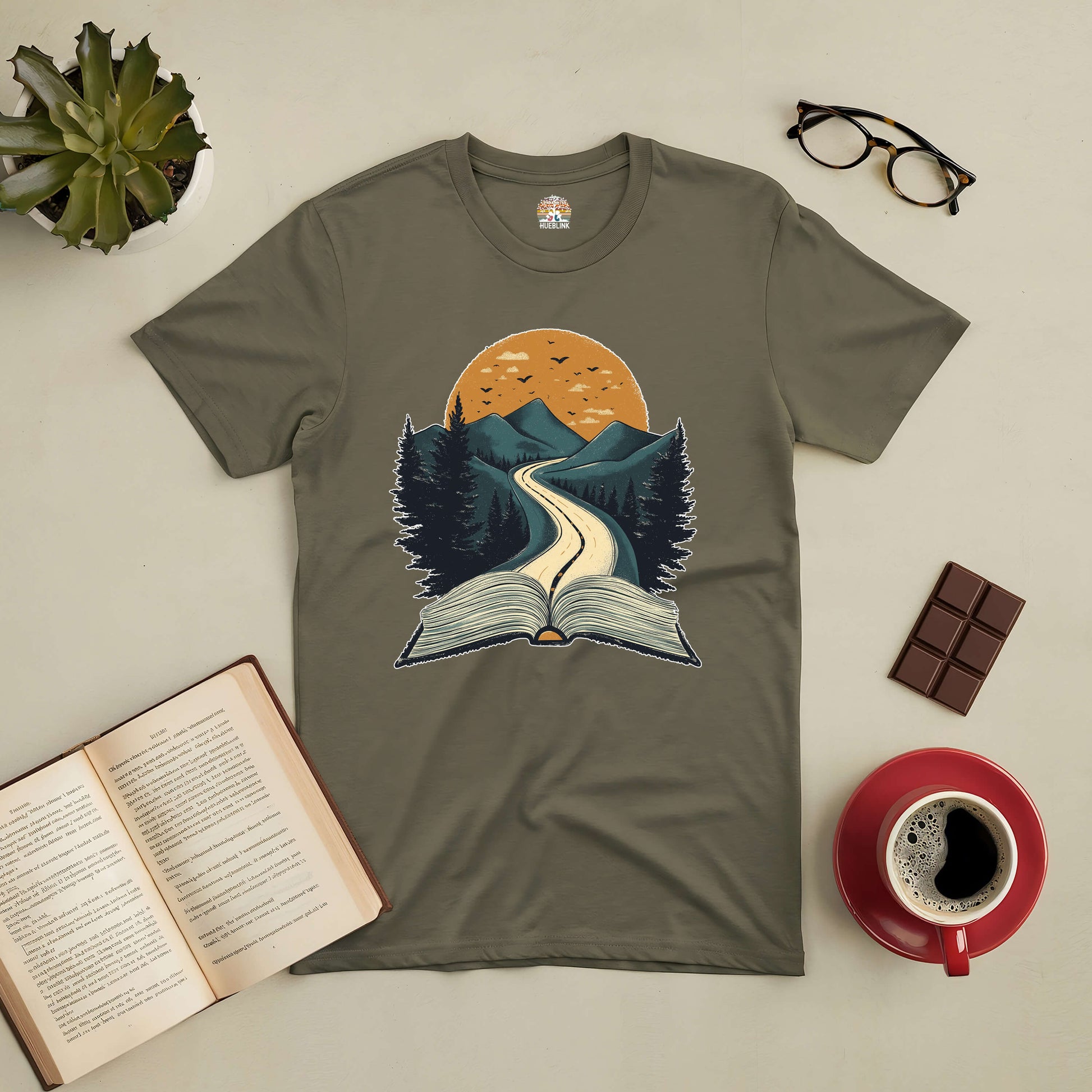 Olive green Starting Point Tee with book and mountain path graphic, surrounded by a book, glasses, coffee, chocolate, and a succulent.