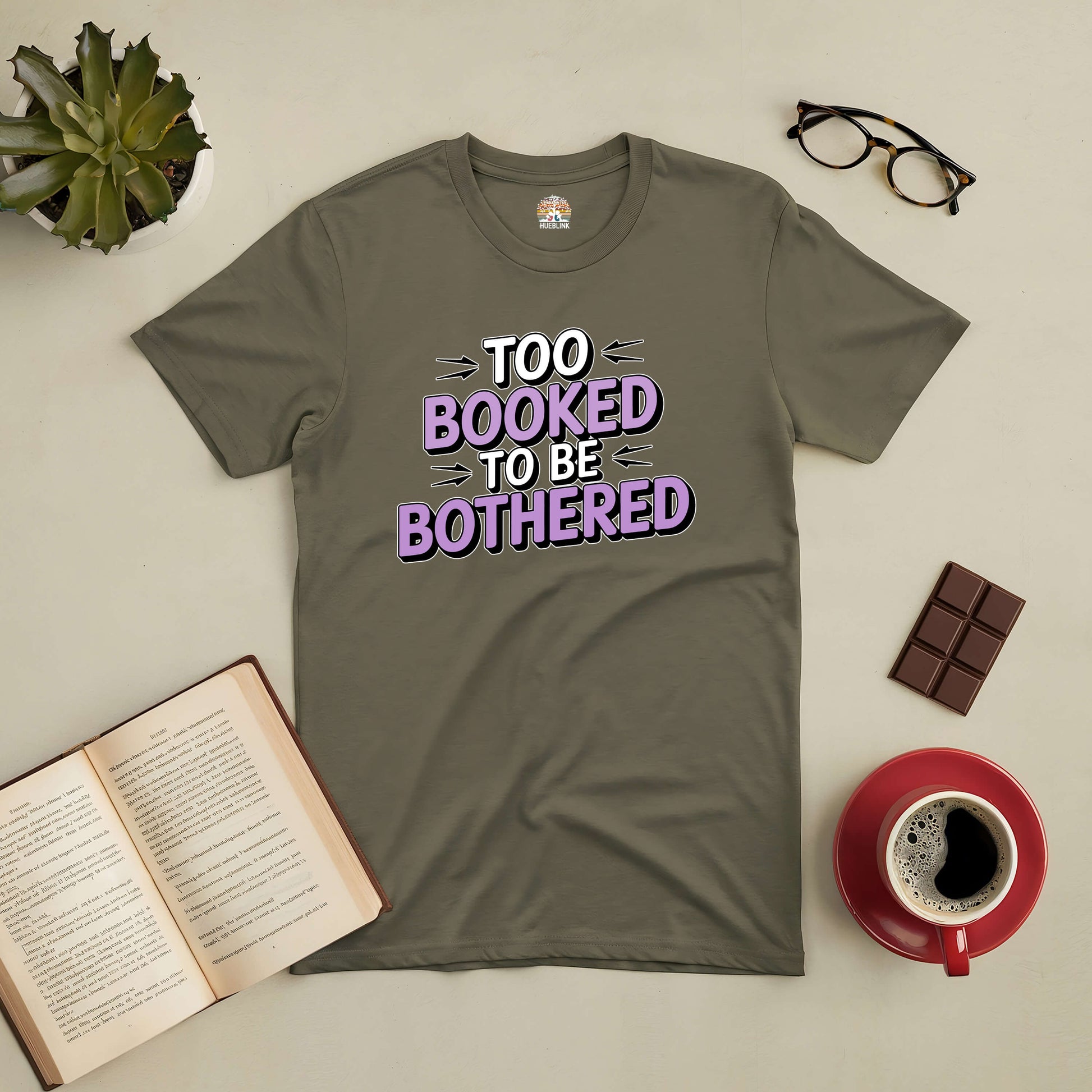 Olive green "Too Booked to Be Bothered" tee laid on a table with an open book, glasses, chocolate, and a cup of coffee.