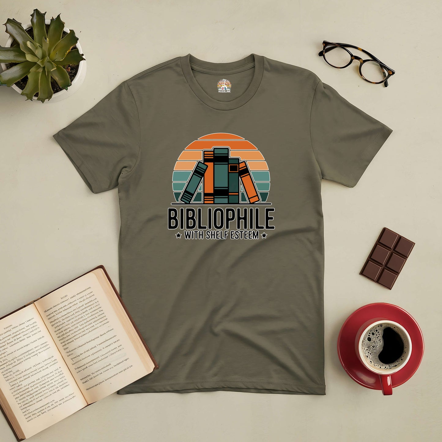 Retro-style "Bibliophile with Shelf Esteem" tee displayed with coffee, book, glasses, chocolate, and plant.