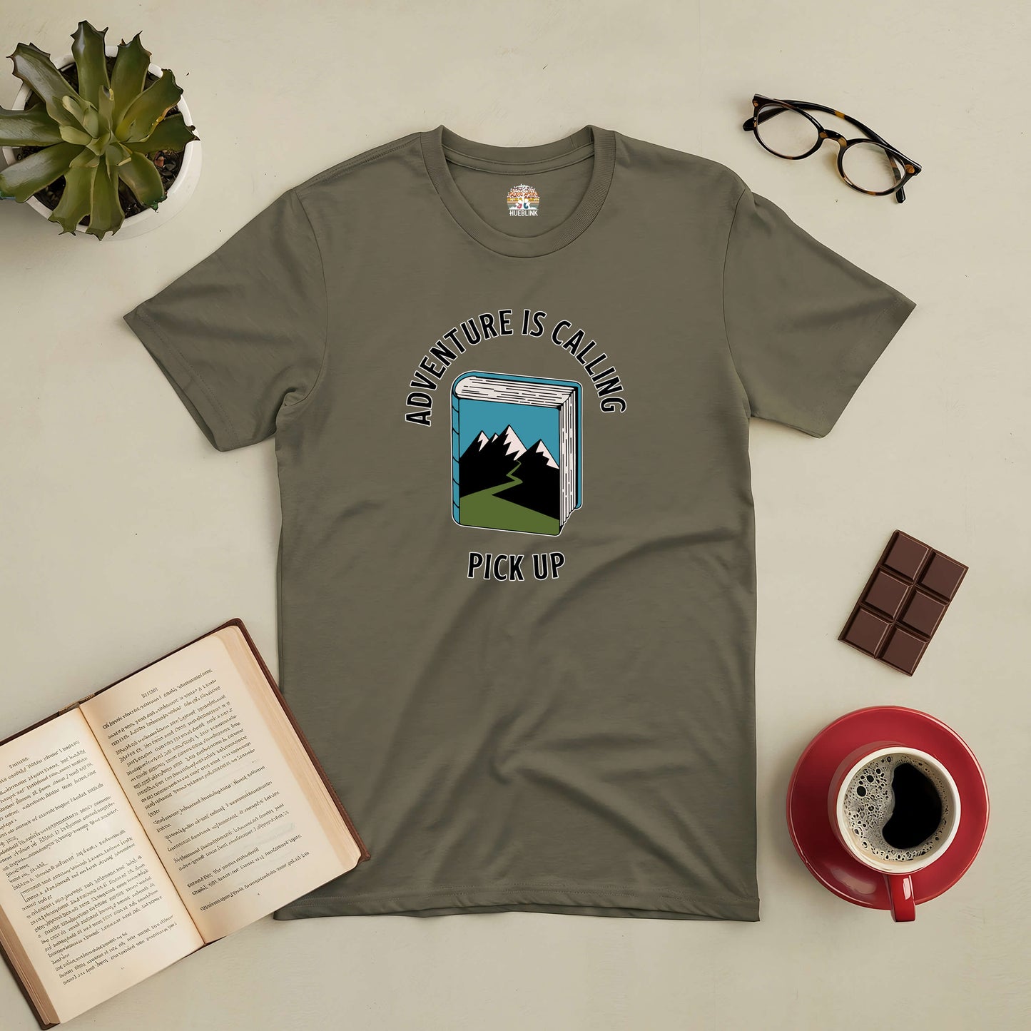 Adventure Is Calling Pick Up Tee with mountain and book design, ideal for explorers, on table with coffee, chocolate, book, and glasses.