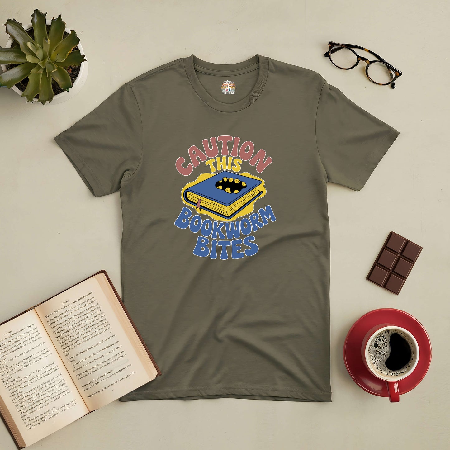 Olive green t-shirt with "Caution, This Bookworm Bites" design, surrounded by glasses, chocolate, coffee, open book, and plant decor.