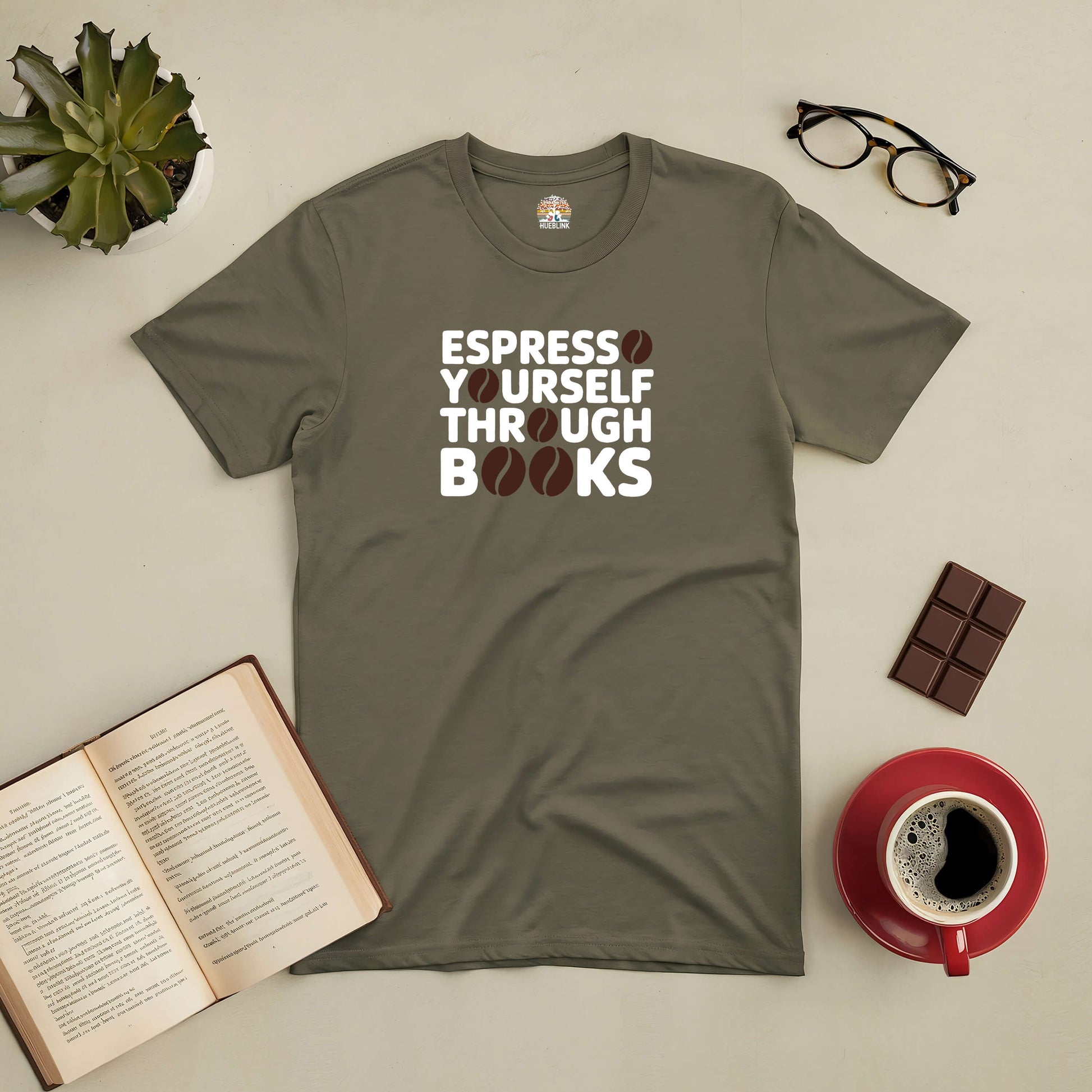 Olive green T-shirt with "Espresso Yourself Through Books" design, surrounded by coffee cup, book, chocolate, glasses, and plant.