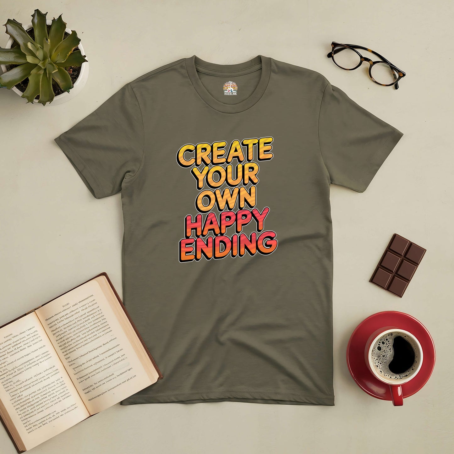 Olive green tee with "Create Your Own Happy Ending" text, surrounded by glasses, chocolate, a book, plant, and a coffee cup.