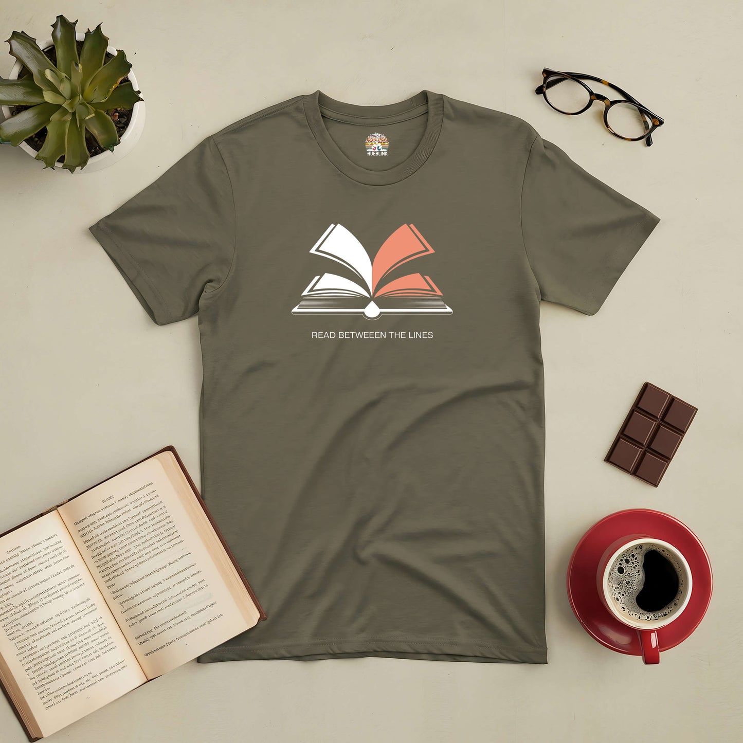 Olive green tee with open book graphic, text "Read Between The Lines", surrounded by glasses, a book, coffee cup, and chocolate.