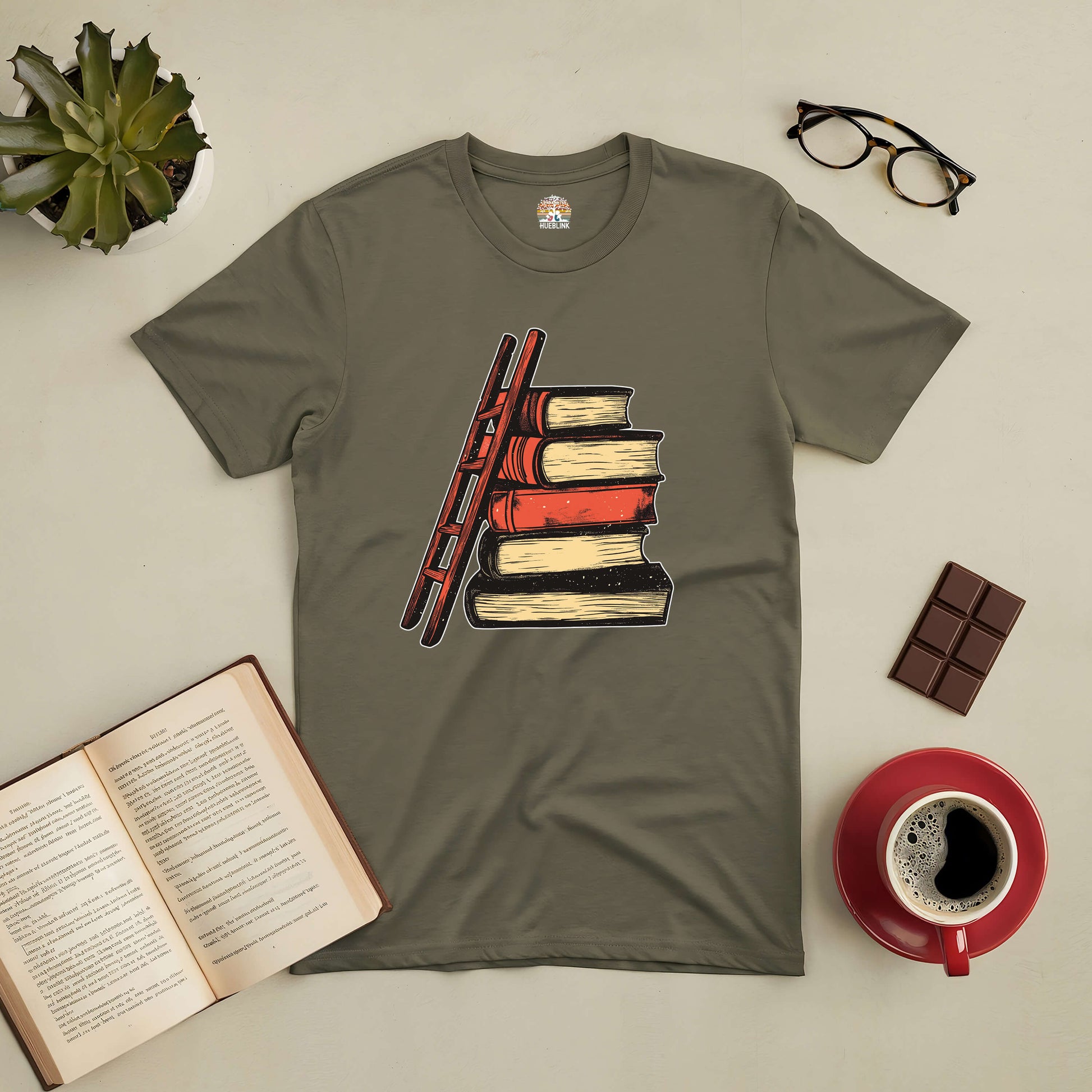 "Reaching New Heights Tee with book and ladder design, surrounded by coffee, chocolate, glasses, open book, and plant"