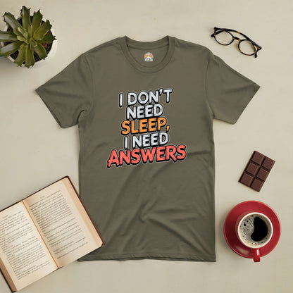 Olive green tee with "I Don't Need Sleep, I Need Answers" text, surrounded by a book, coffee, glasses, and chocolate on a table.