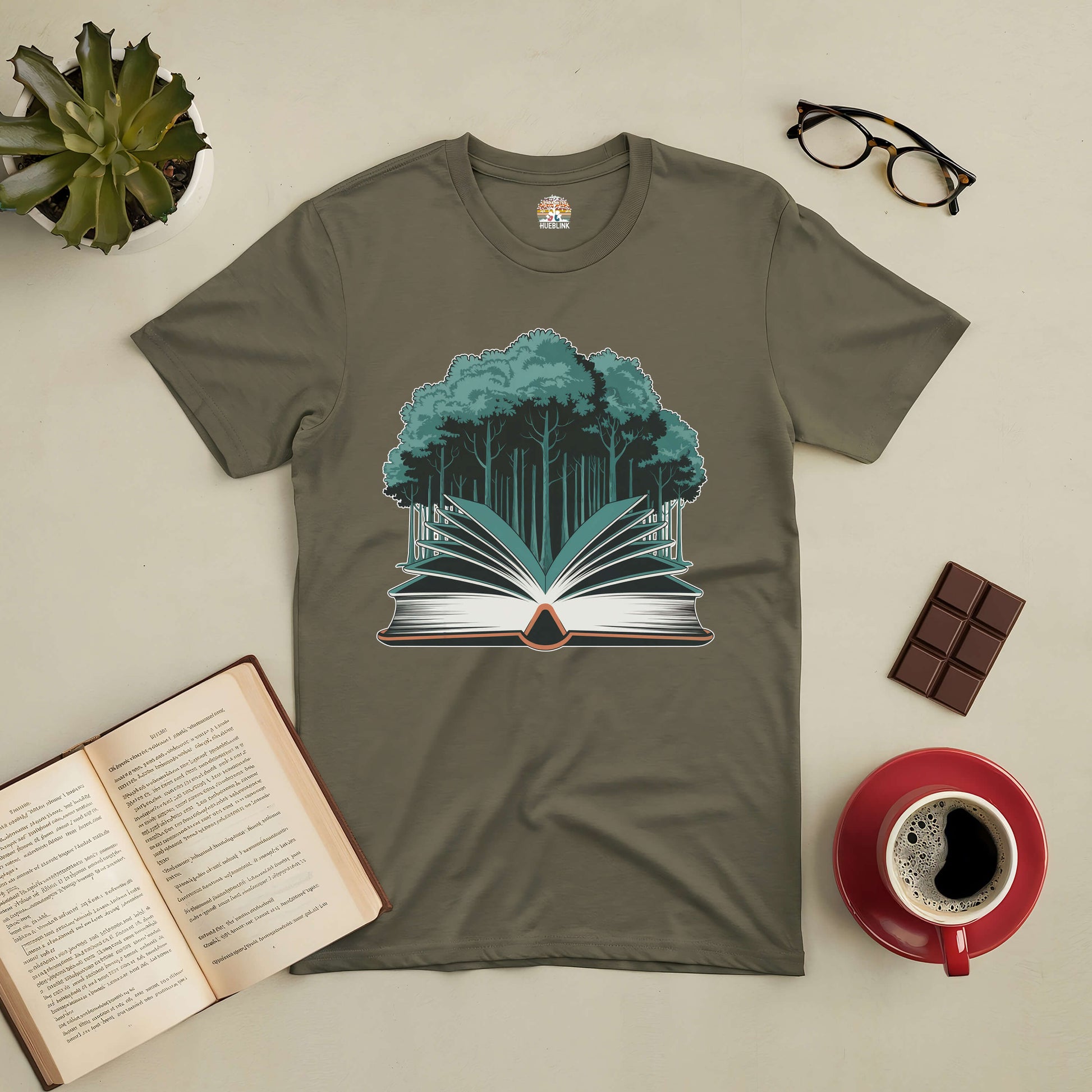 Rooted in Story Tee featuring forest and book design, surrounded by an open book, glasses, chocolate, coffee, and a plant.