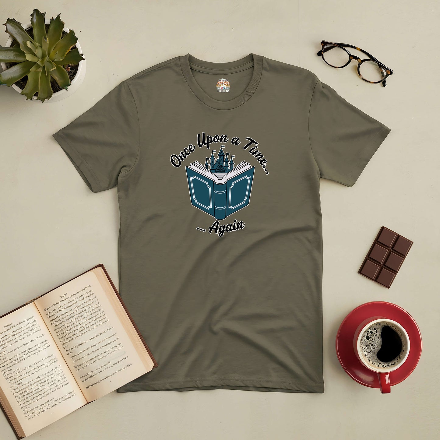 Olive green "Once Upon a Time... Again" tee with fairy tale design, surrounded by open book, coffee cup, chocolate, and glasses.