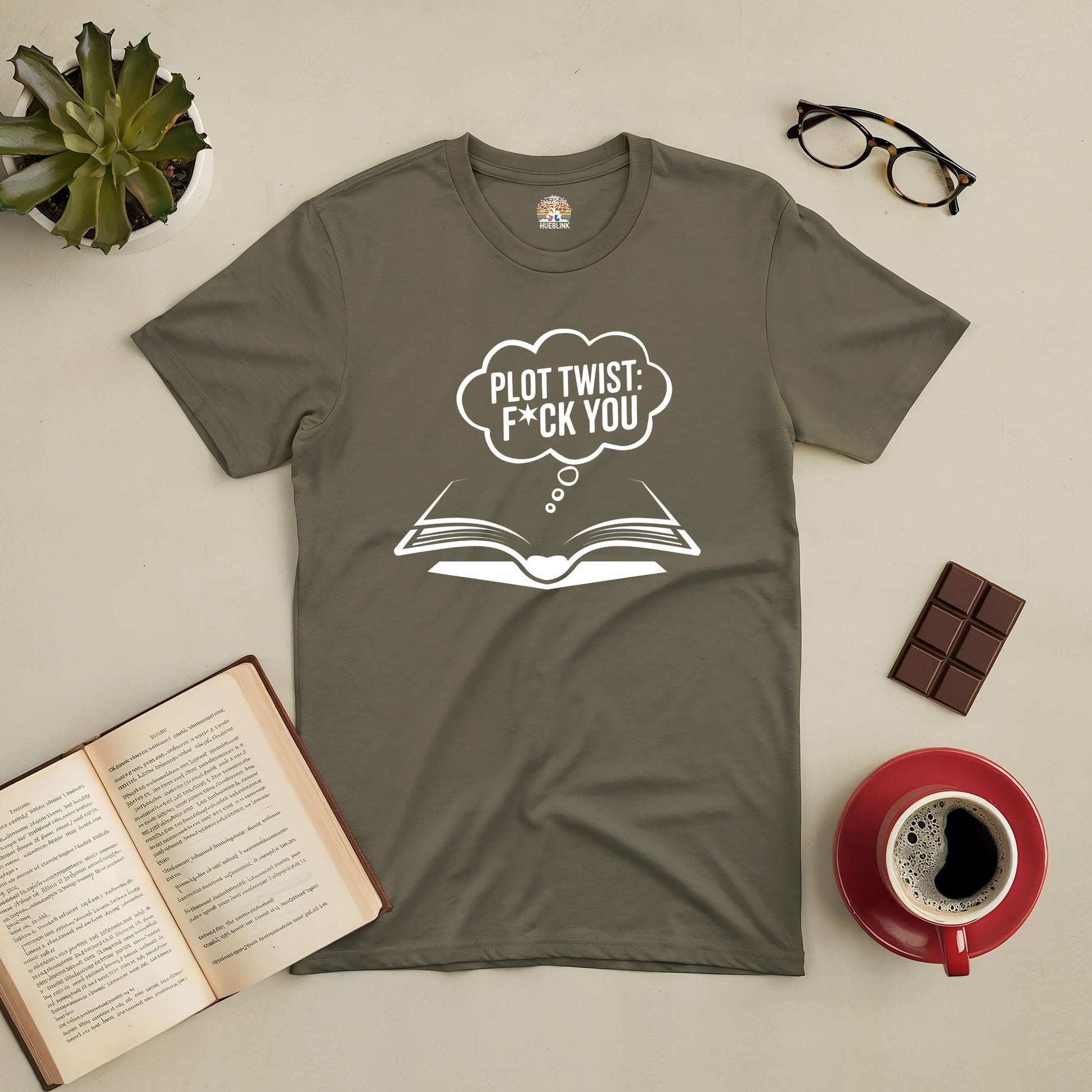 Olive green "Plot Twist: F*ck You" t-shirt displayed with books, glasses, a plant, chocolate, and a coffee cup on a flat surface.