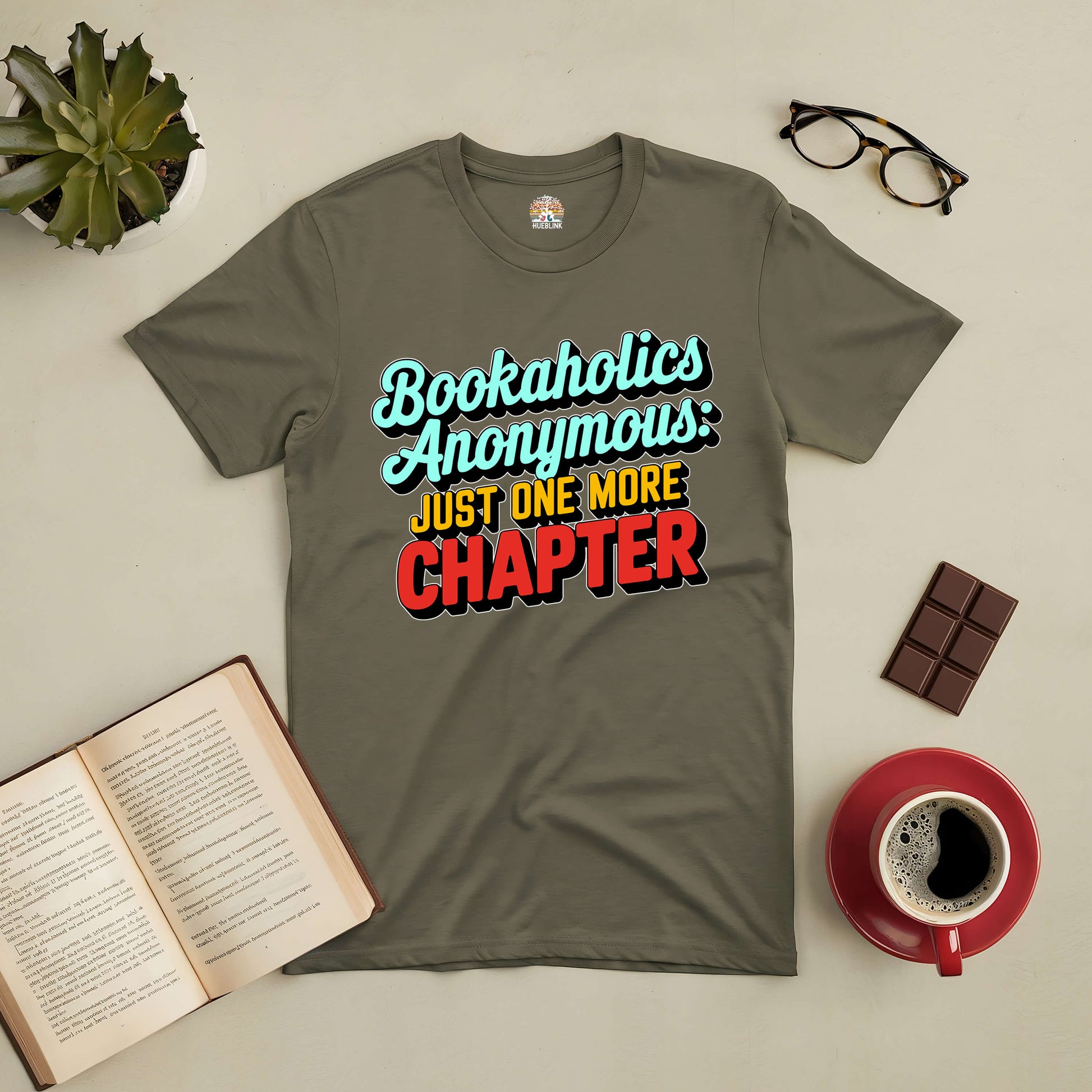 Bookaholics Anonymous Tee with "Just One More Chapter" text, perfect for readers, surrounded by a book, coffee, and chocolate.