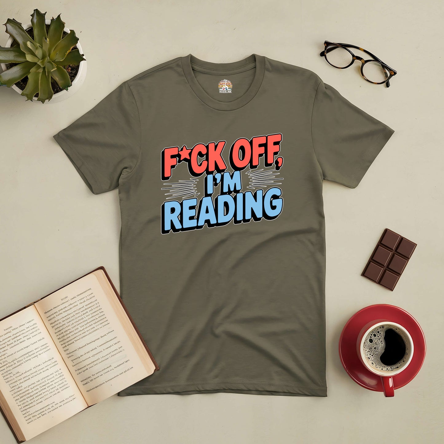 "Bold Tee with 'F*ck Off, I'm Reading' message on a table with open book, chocolate, glasses, coffee, and plant"