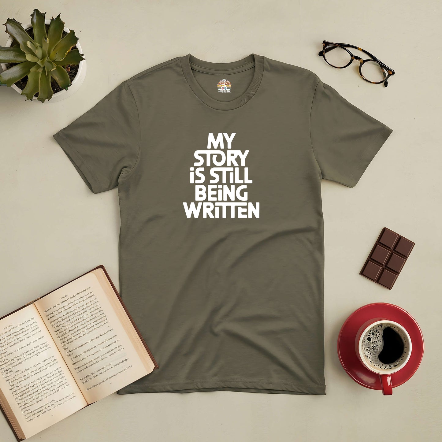 Olive green "My Story Is Still Being Written" tee beside an open book, coffee, and glasses on a table.