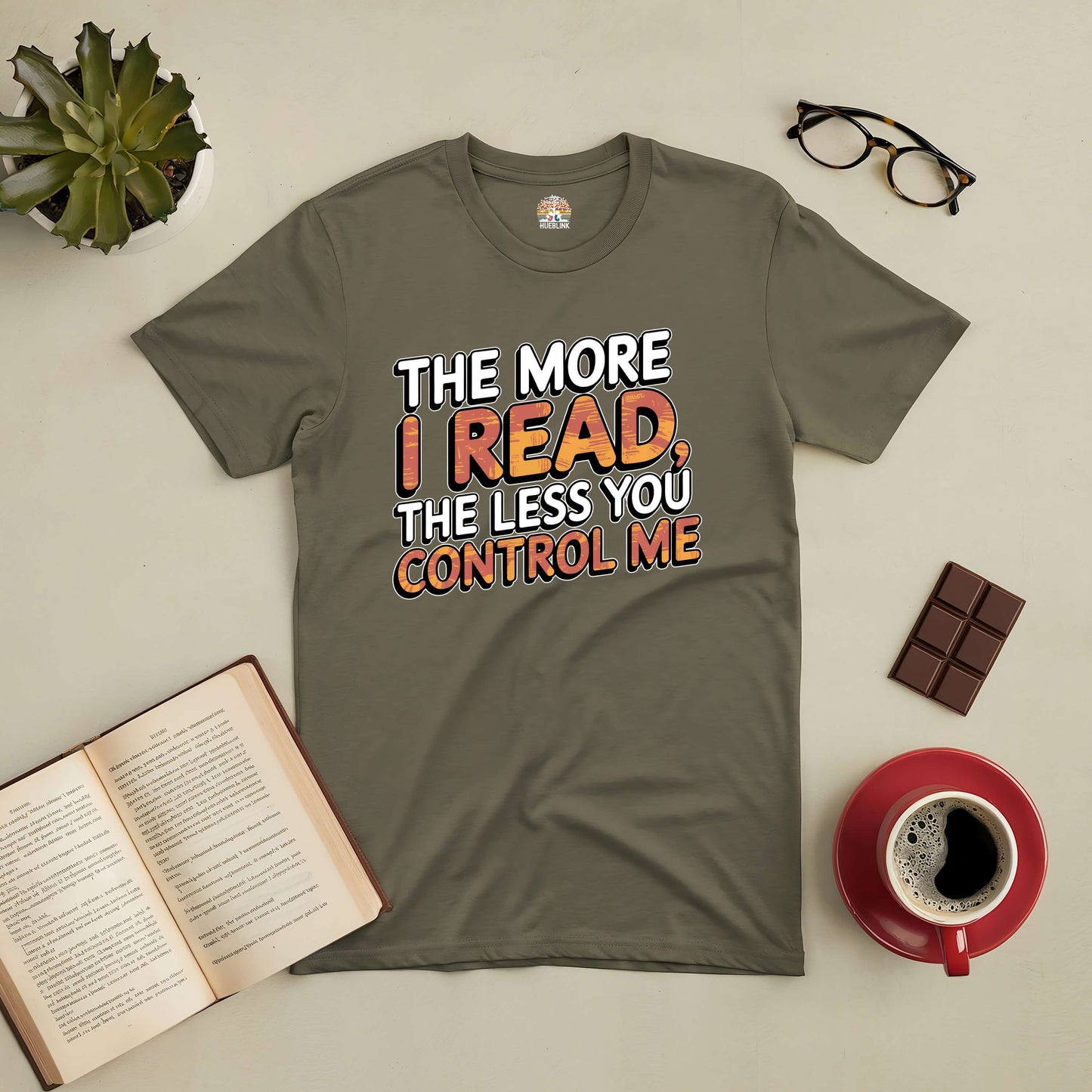 Olive green tee with "The More I Read, The Less You Control Me" text, surrounded by a book, glasses, chocolate, and coffee.